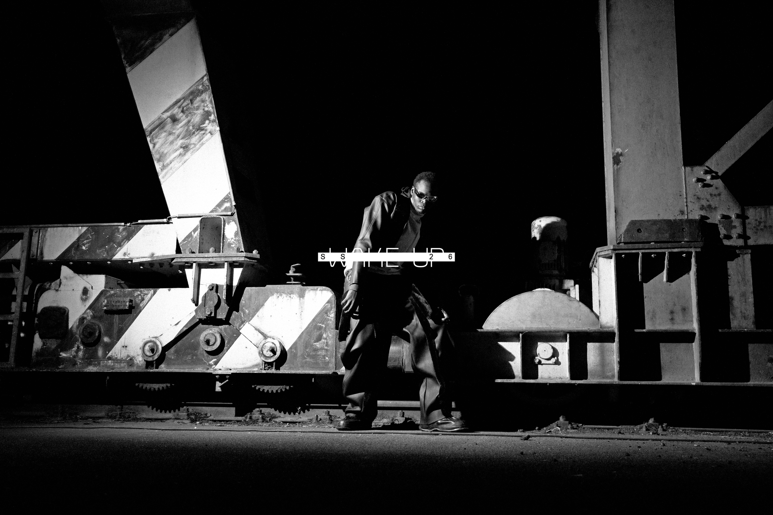 A person wearing sunglasses and a jacket, standing and leaning on a large industrial piece of equipment in a dark setting. Daniel Alexander Weiter - DAW