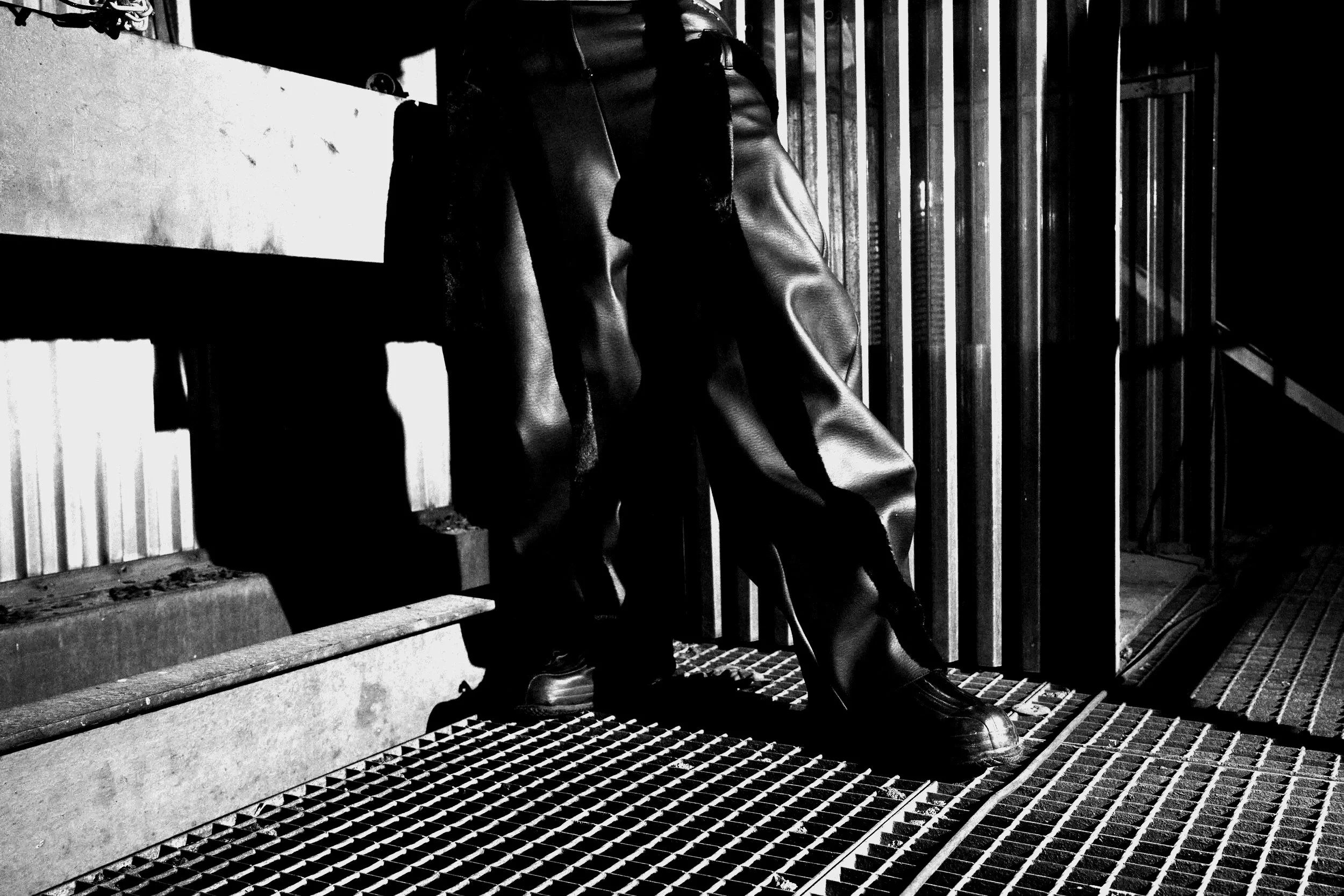 A person sitting on steps beside a metal railing, wearing a leather pants and boots, with a barred fence and concrete wall in the background. Daniel Alexander Weiter - DAW