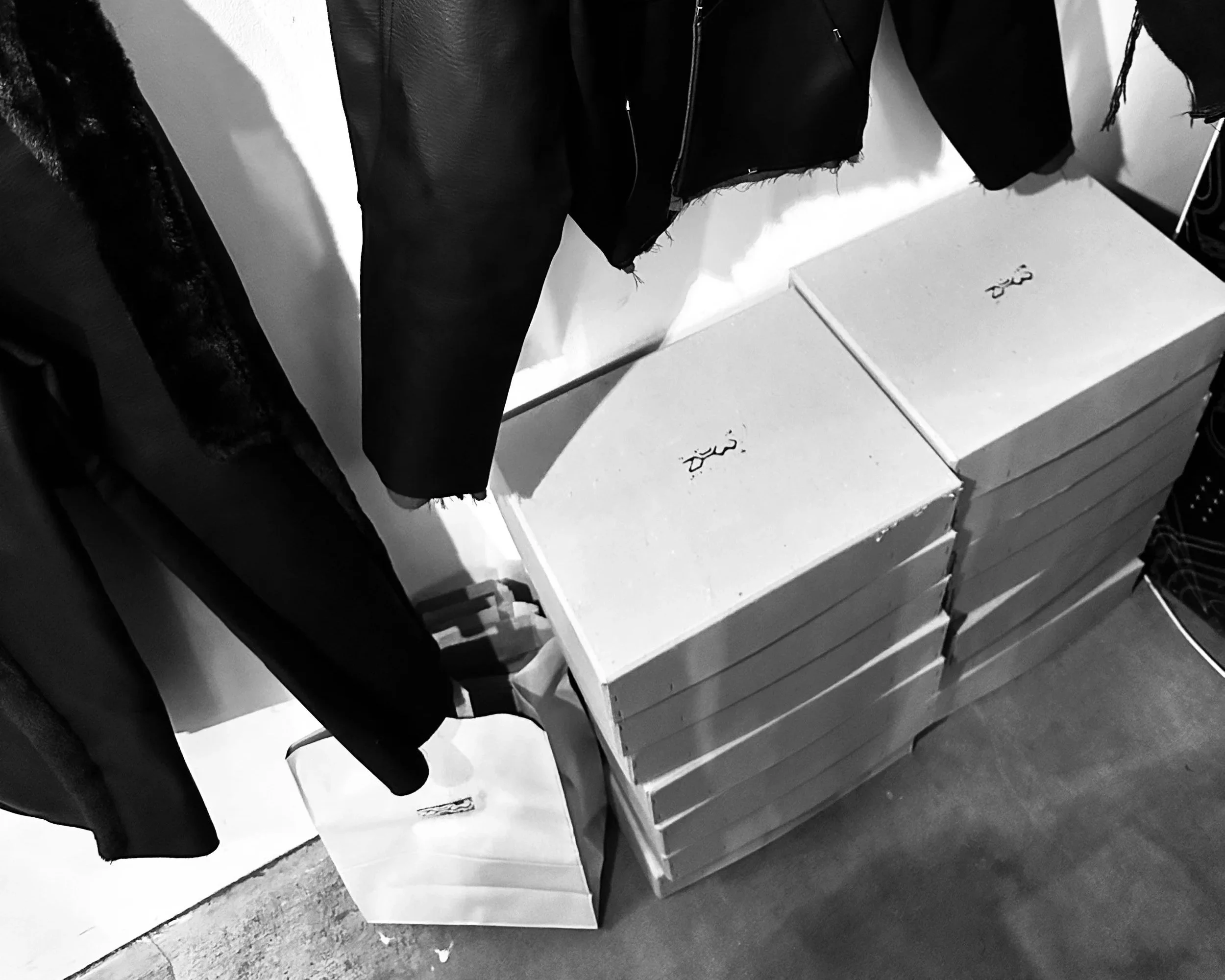 Black and white photo showing a row of stacked cardboard boxes on a floor next to white shelves, with dark clothing hanging on the wall. Daniel Alexander Weiter - DAW
