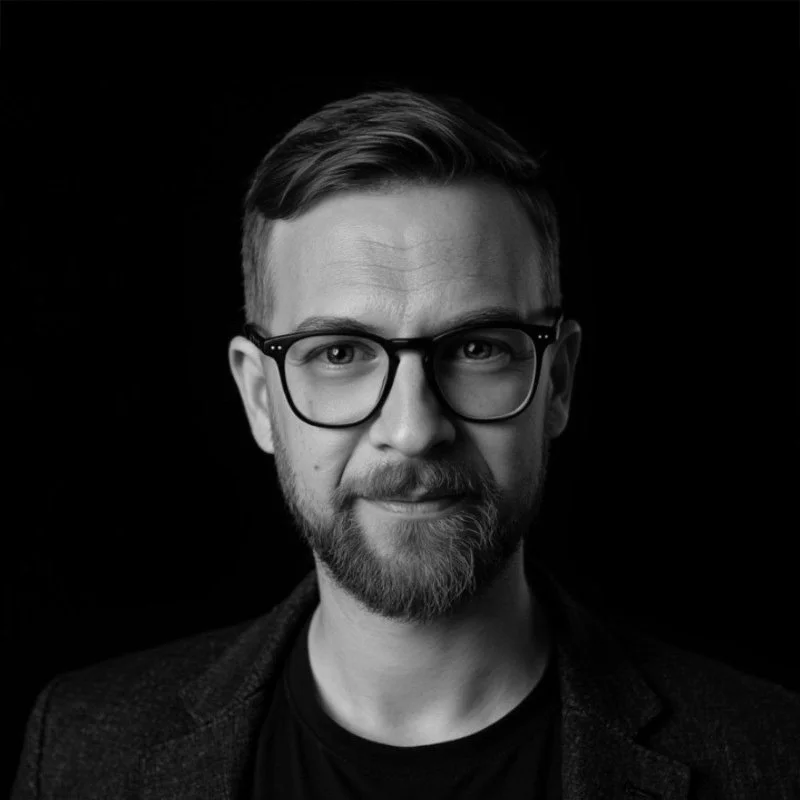 Black and white portrait of a man with glasses, a beard, and short hair, wearing a dark blazer and shirt, against a dark background.