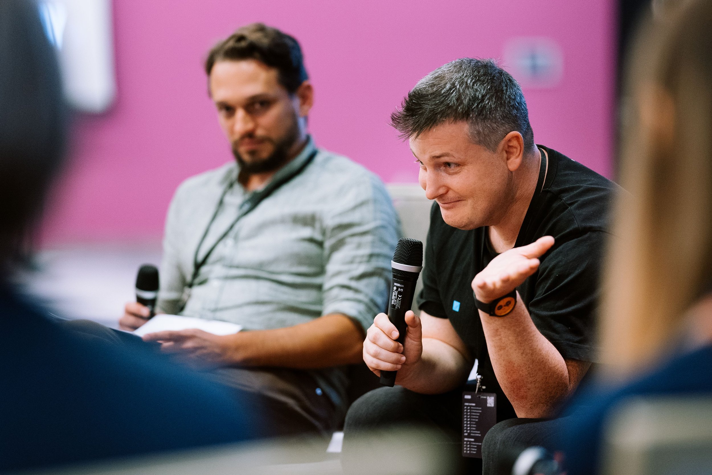 Two men sitting, one speaking into a microphone, in a discussion panel setting