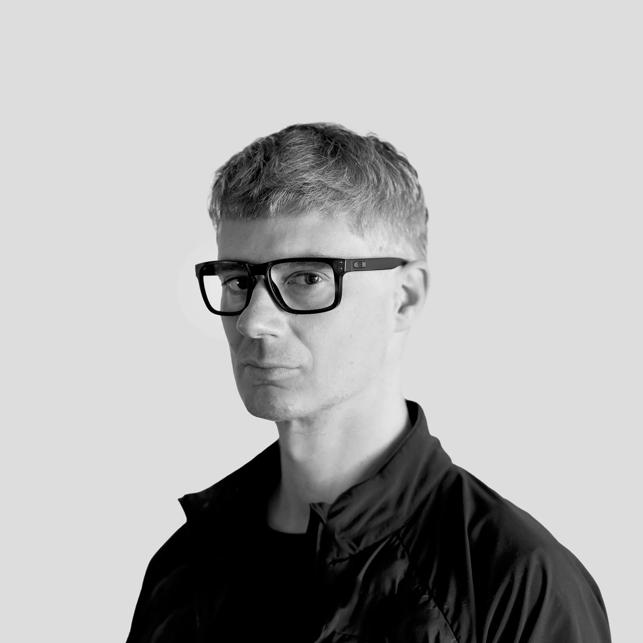 Black and white portrait of a man with short, light hair wearing thick black glasses and a black jacket, looking slightly to the side against a plain background.