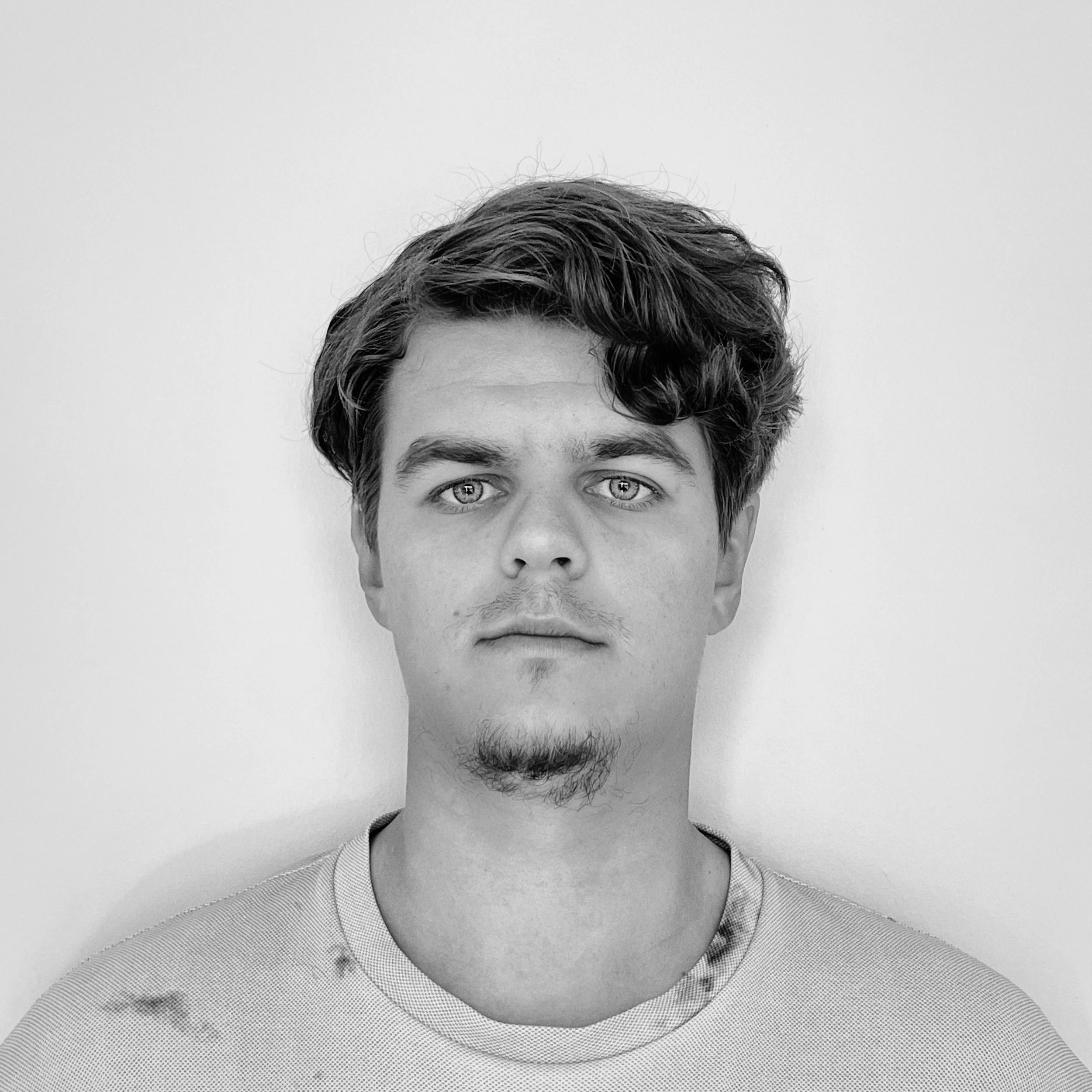 A young man with short, wavy hair and light eyes looking directly at the camera, wearing a light-colored shirt, against a plain background.