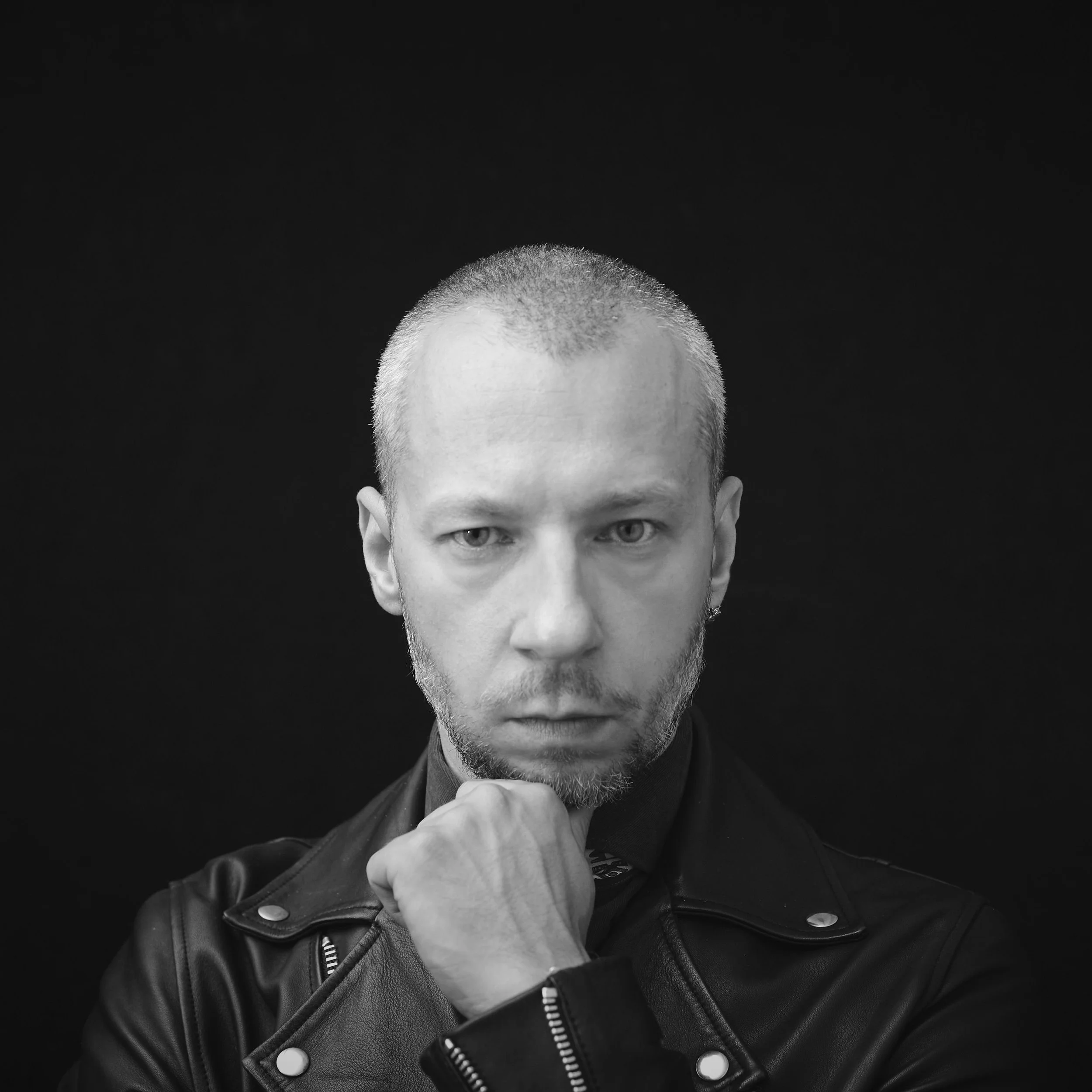 Black and white portrait of a man with a shaved head and beard, wearing a leather jacket, looking directly at the camera with his hand under his chin, against a black background.