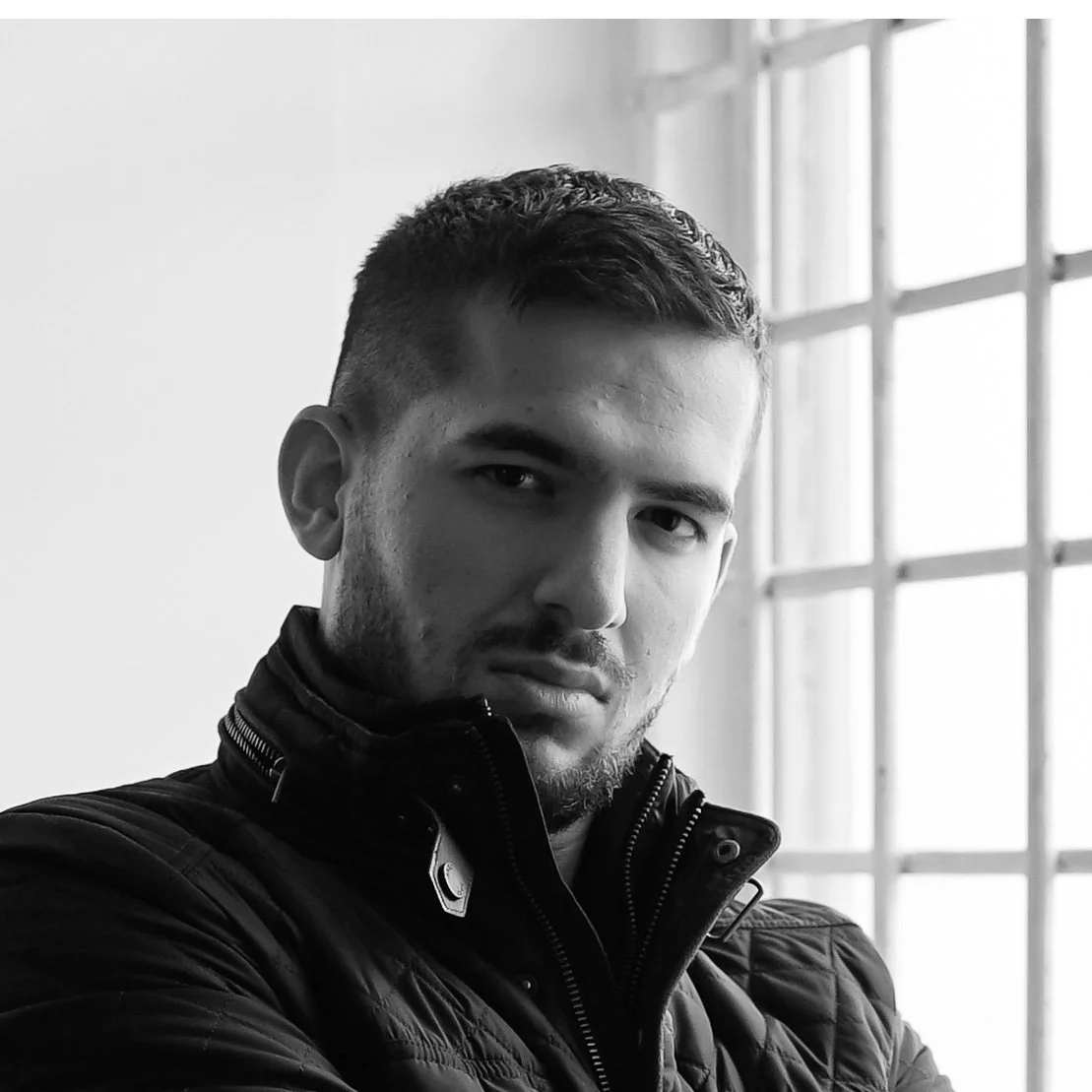 Black and white portrait of a young man with short dark hair and a beard, wearing a black jacket, standing in front of a window with a grid pattern.