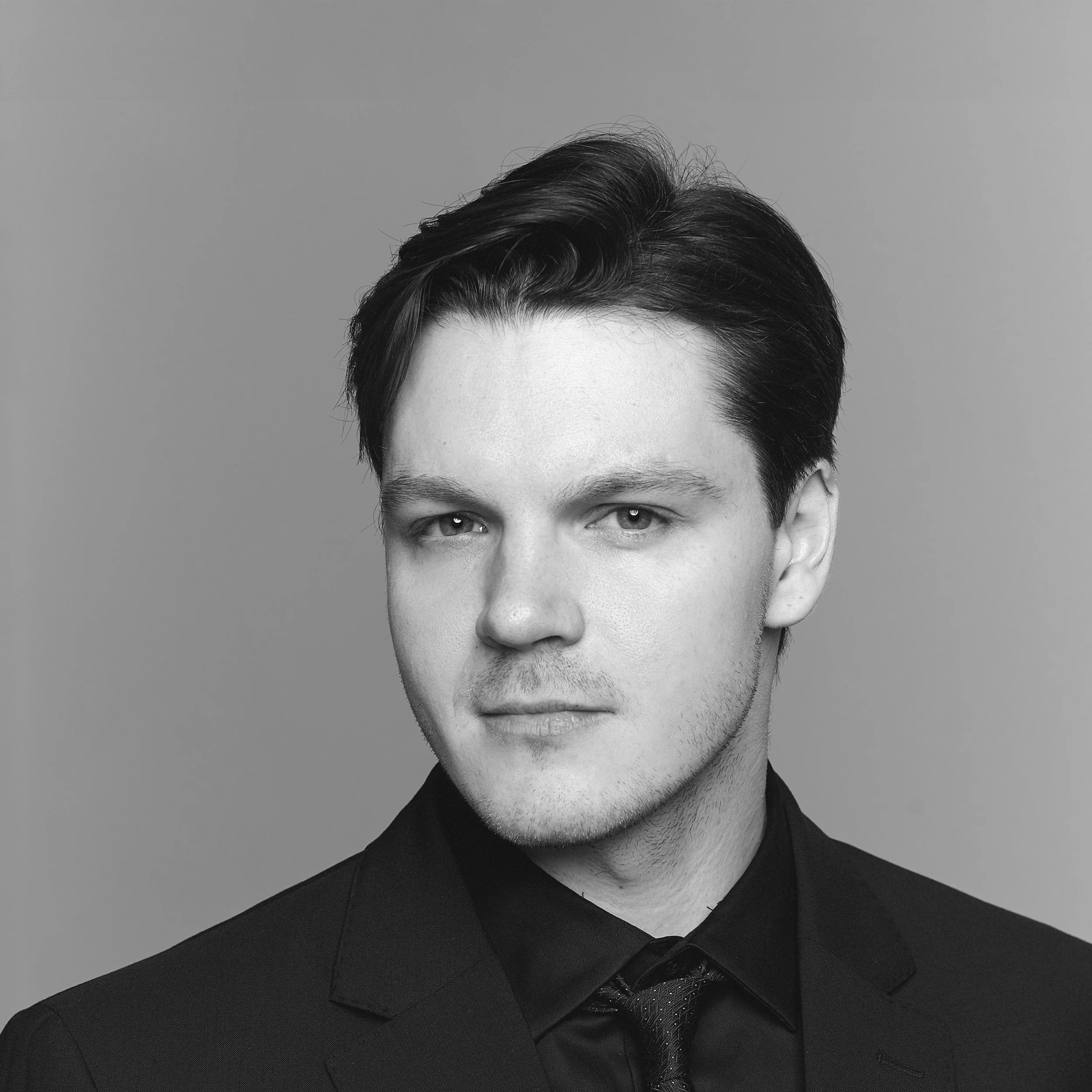 Black and white portrait of a young man with dark hair, wearing a suit and tie, looking directly at the camera.