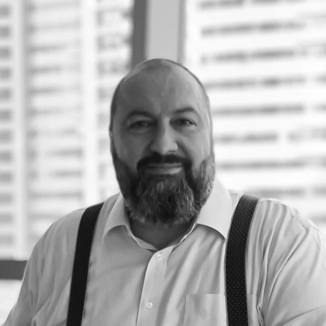 Black and white photo of a man with a beard, wearing a white shirt and suspenders, smiling at the camera indoors in front of window blinds.