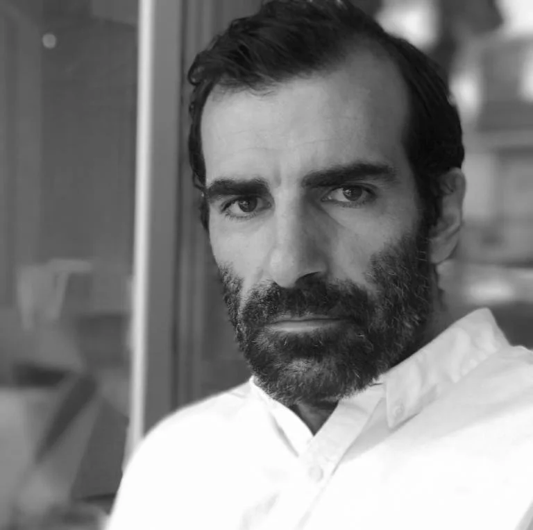 Black and white close-up photo of a man with dark hair and a beard, wearing a collared shirt, looking directly at the camera.