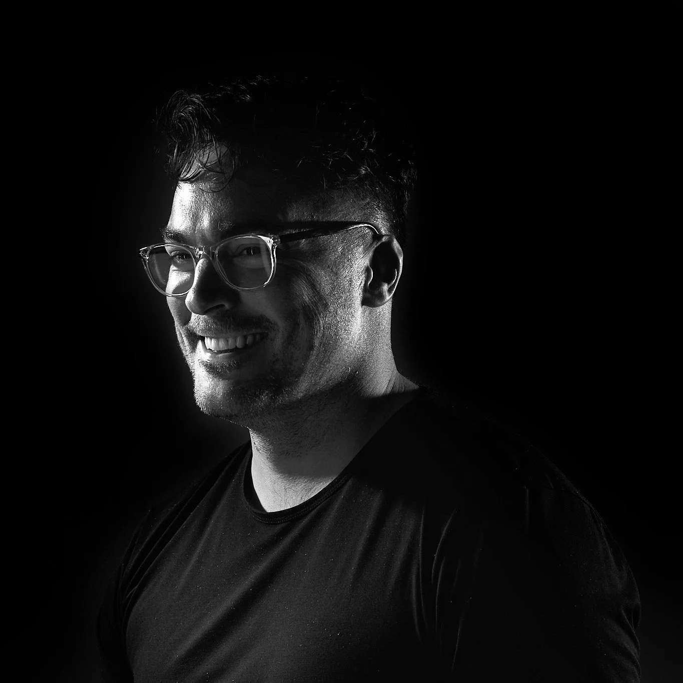 Black and white portrait of a smiling man wearing glasses, with short wavy hair, wearing a black shirt, against a dark background.