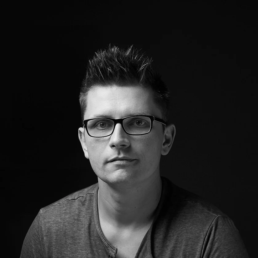 Black and white portrait of a young man with glasses, short spiked hair, wearing a t-shirt, against a dark background.