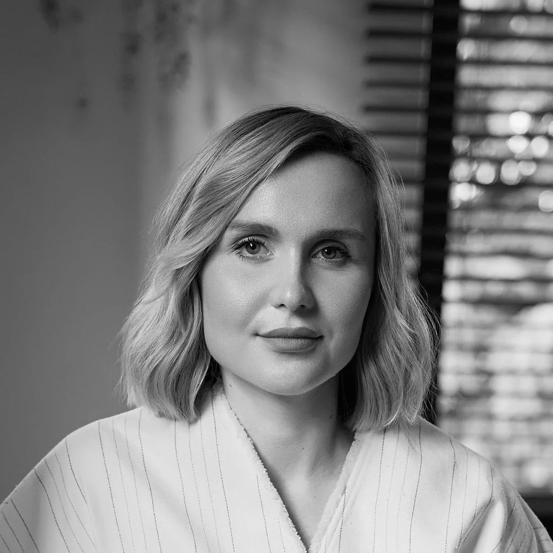 Black and white portrait of a woman with shoulder-length wavy hair, wearing a light-colored top, standing near window blinds.
