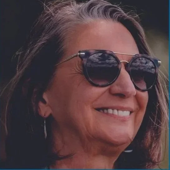 Une femme souriante portant des lunettes de soleil, cheveux bruns, à l'extérieur.