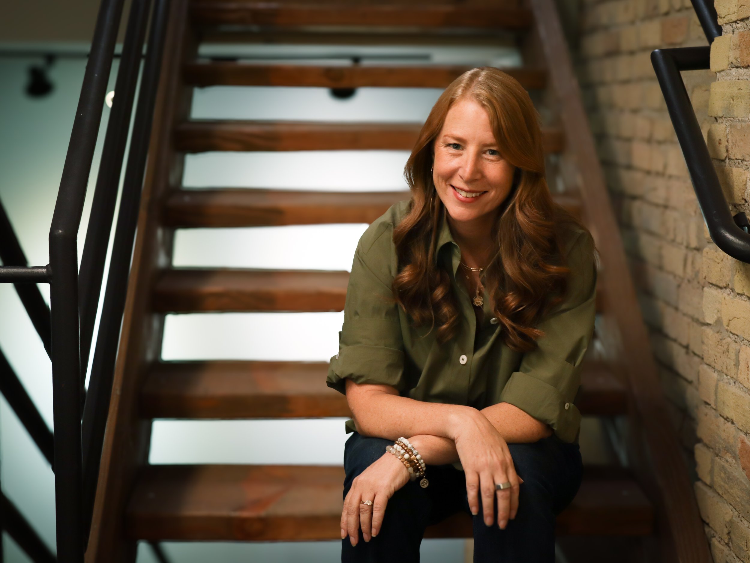A smiling woman with long red hair sitting on a staircase, resting her arms on her knees, wearing an olive green shirt and beaded bracelets, with a brick wall on her right side.