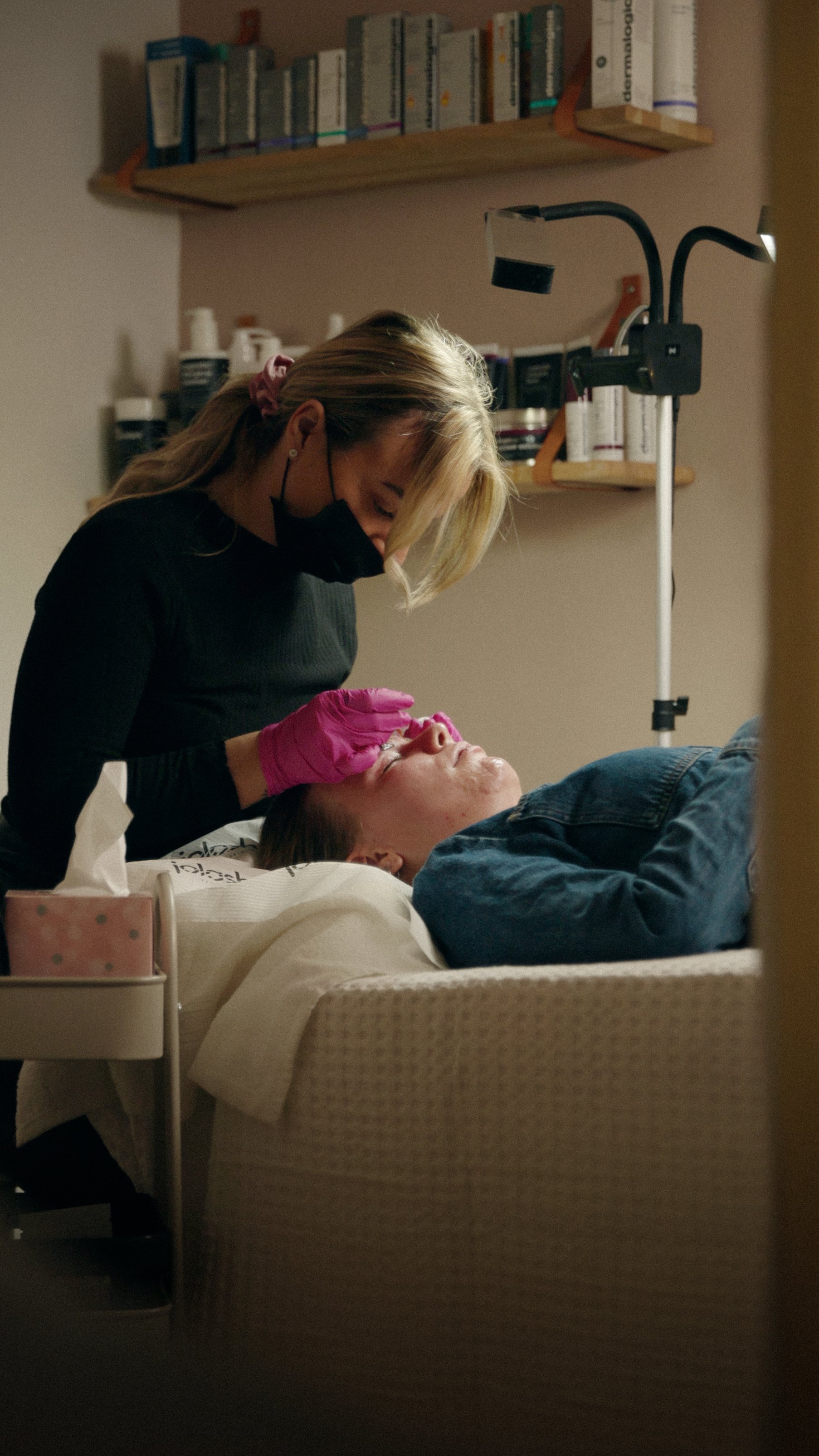 En sygeplejerske, der iført pink handsker og sort ansigtmaske, undersøger en patients øje på et hospital eller klinik.