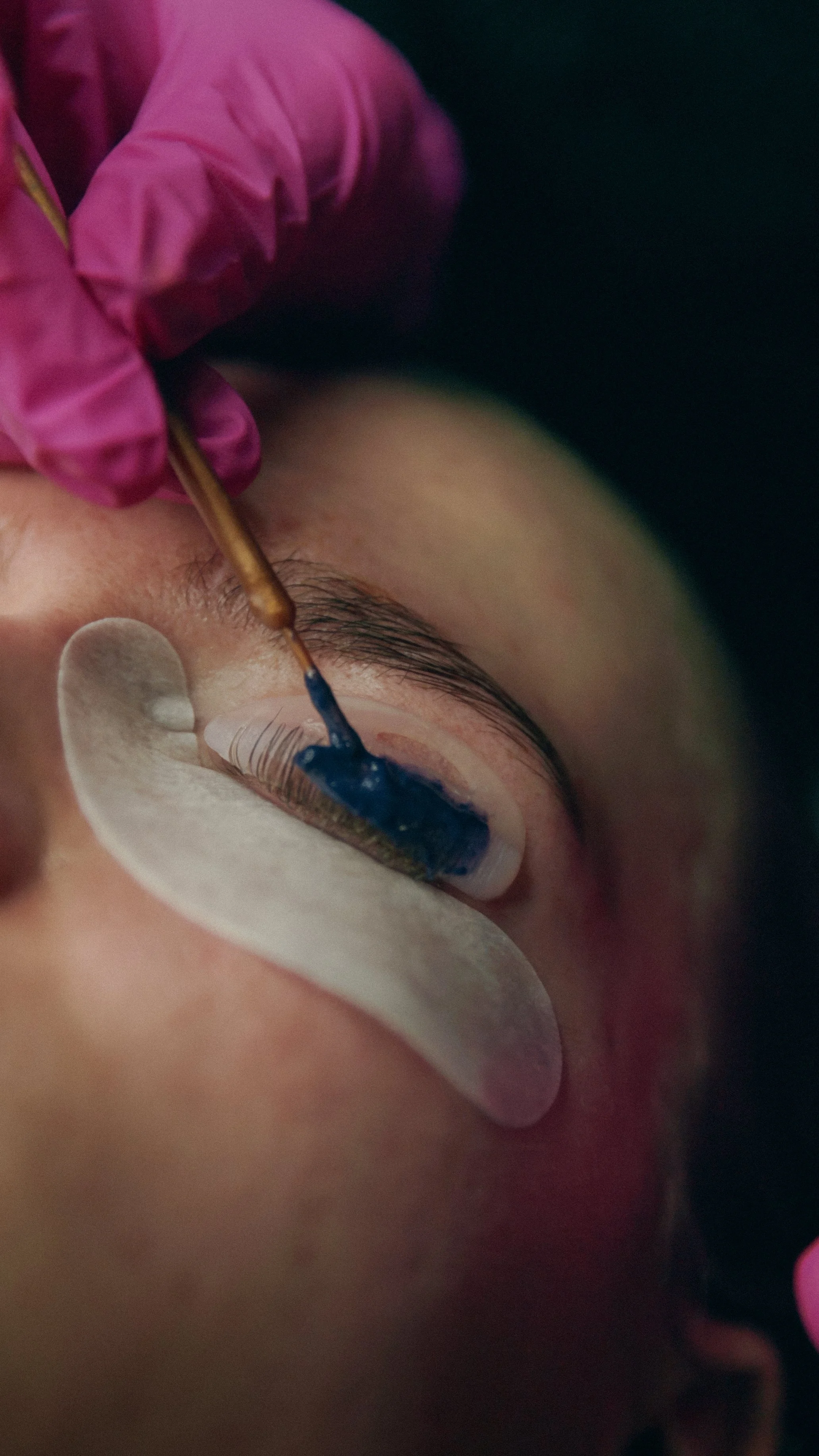 En person får lavet en permanent øjenbryn tatovering ved at påføre blå farve på øjenbrynene med en kosmetisk nål, mens en anden person bruger en pink glovet hånd til at håndtere værktøjet.