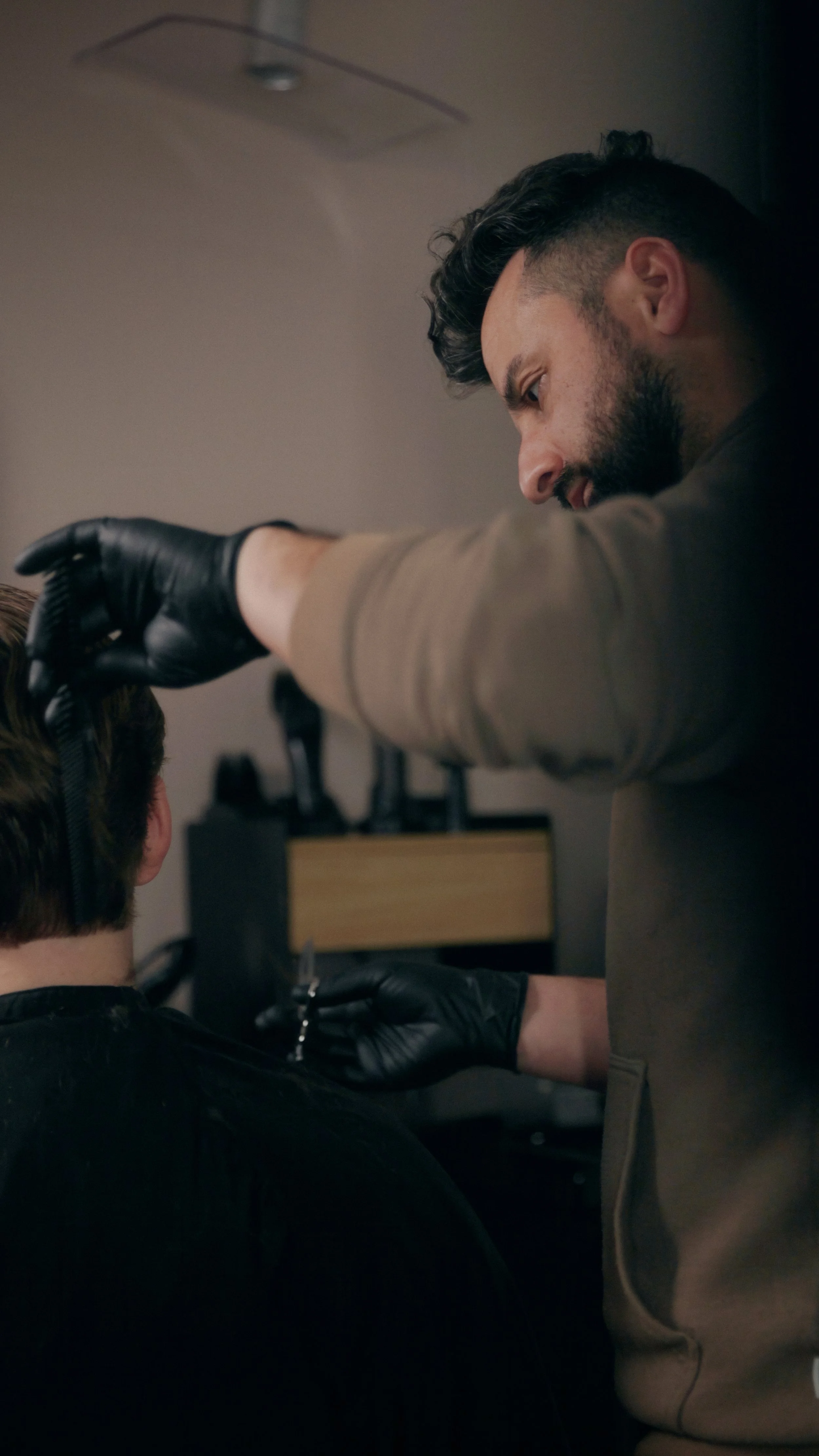 En mand med sort hår og skæg, iført grå sweater, arbejder med en person, der har brunt hår, i en setting, der ligner en frisørsalon, iført sort tøj, med sort hår og hænder i sorte handsker, mens han bruger en saks.