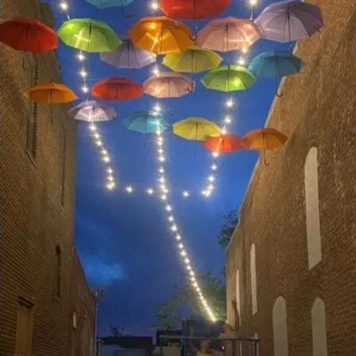 Umbrellas hanging upside down in the sky between buildings with string lights at night.
