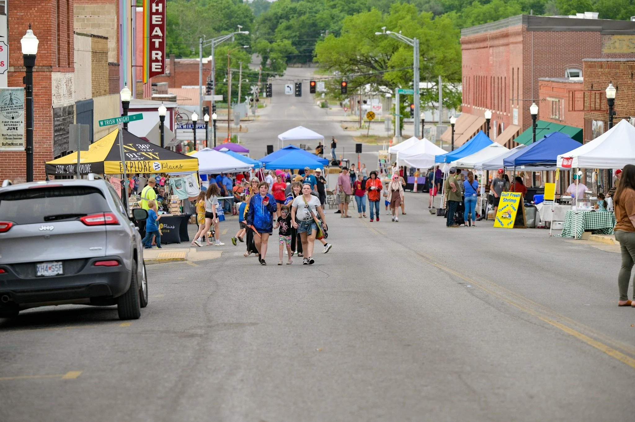 A small town street with street vendors selling food and crafts under tents, with people walking and browsing, and a traffic light in the distance.