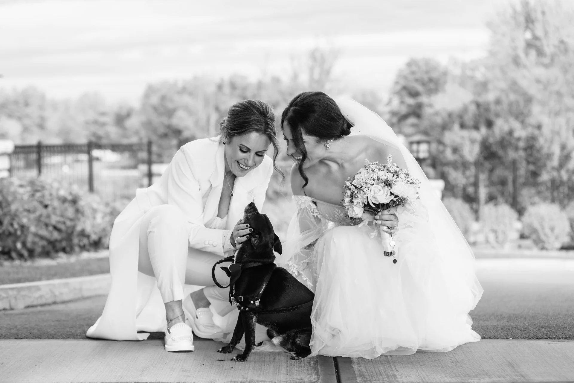 Two women, one in a wedding dress holding a bouquet, and the other in white suspenders and pants, smiling and petting a small black dog outdoors, with trees and a fence in the background.
