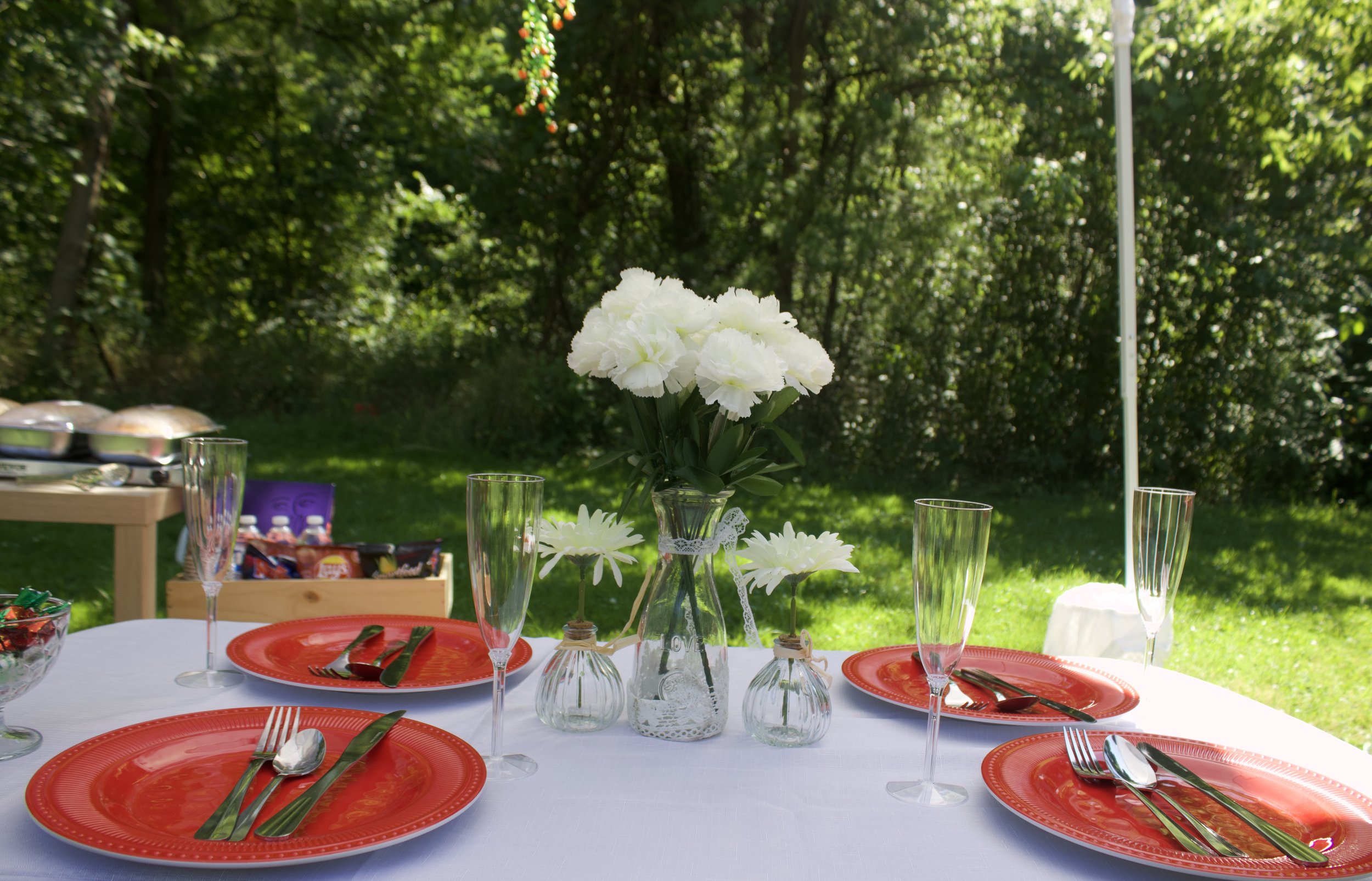 Outdoor table setting with four red plates, silverware, and four champagne flutes, centered around a vase of white flowers, with a lush green garden in the background.