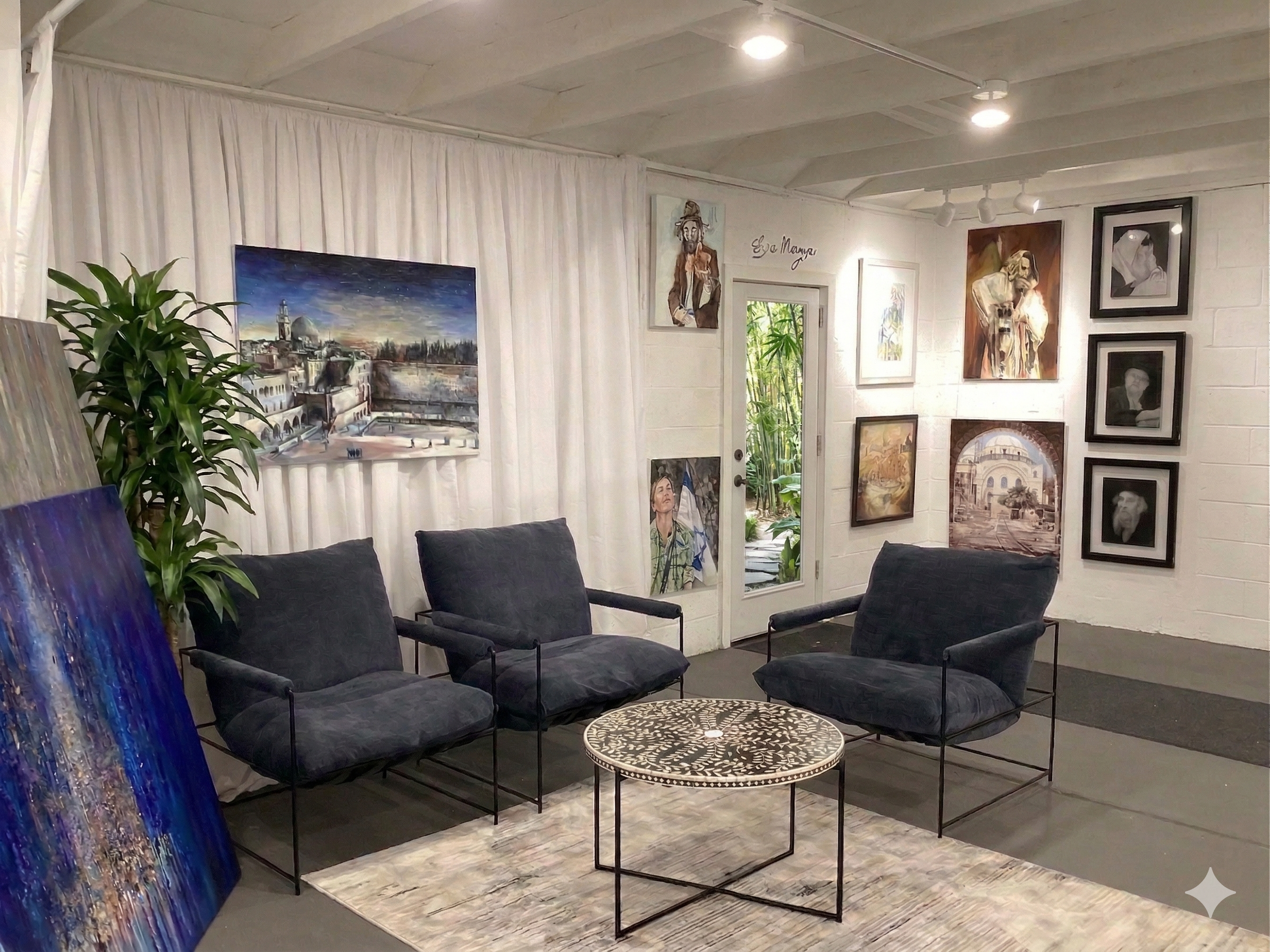 with four black chairs, a round patterned coffee table, and multiple paintings on white walls. There is a door with a glass panel showing greenery outside, and paintings include a cityscape, figures, and portraits. A large plant is next to the chairs, and a large abstract painting rests against the wall.