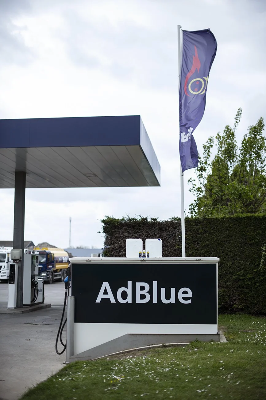 Signage bij een tankstation met de tekst "AdBlue". Een vlag met een logo of ontwerp wappert boven het station, en er is een pompstation zichtbaar op de achtergrond.
