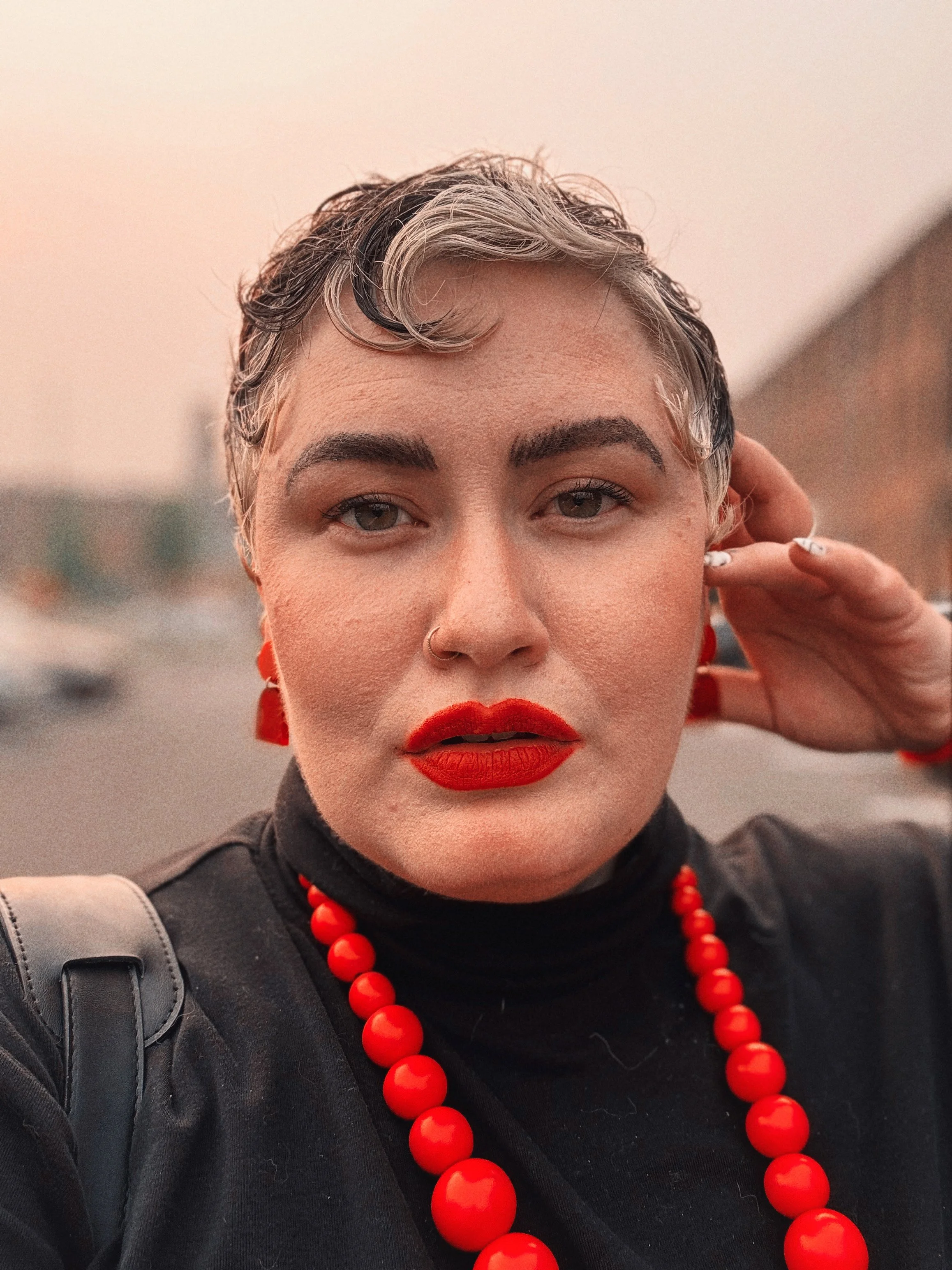 Photo of a feminine person with short curly hair. They have silvery blond highlights and red lips. They're wearing a black turtleneck and red jewelry.