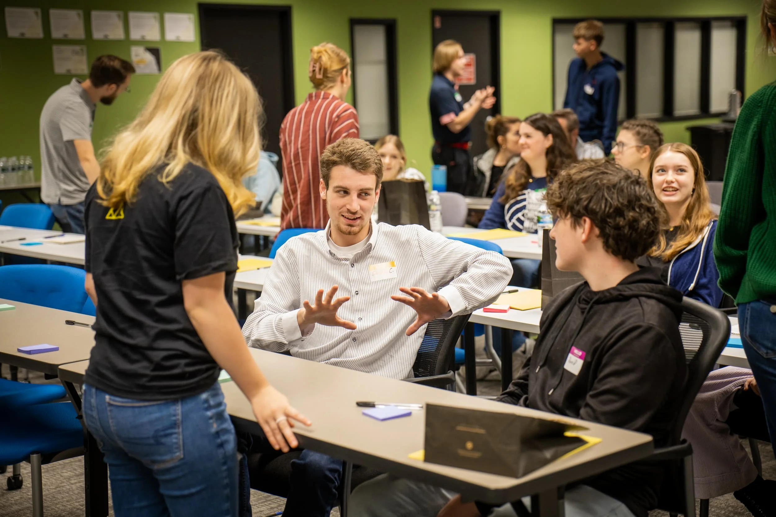 Group of people engaging in conversation during a workshop or meeting in a conference room with green walls and windows.