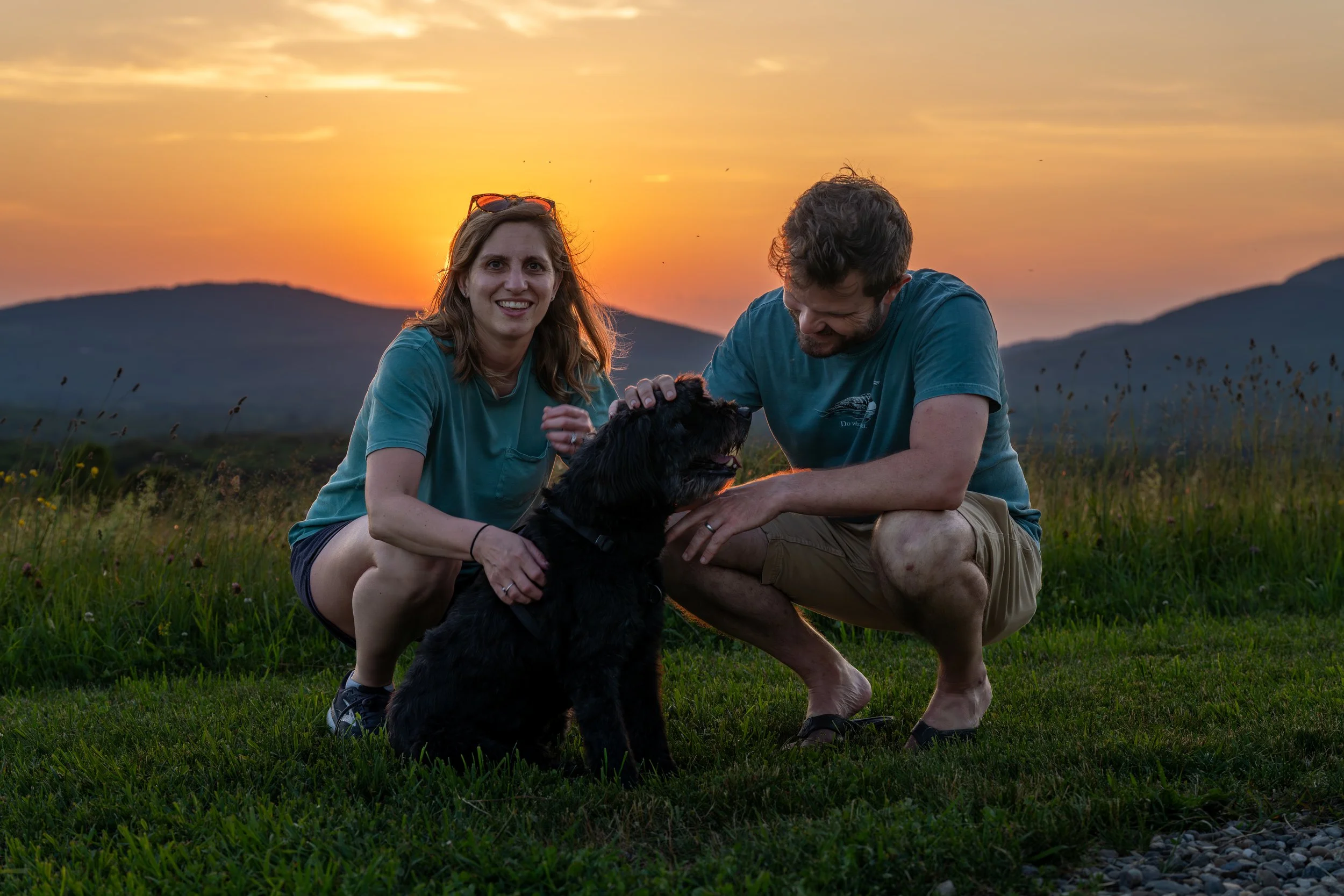 Couple with their black dog at sunset at Beech Hill Preserve in Rockport, Maine, with mountains in the background.