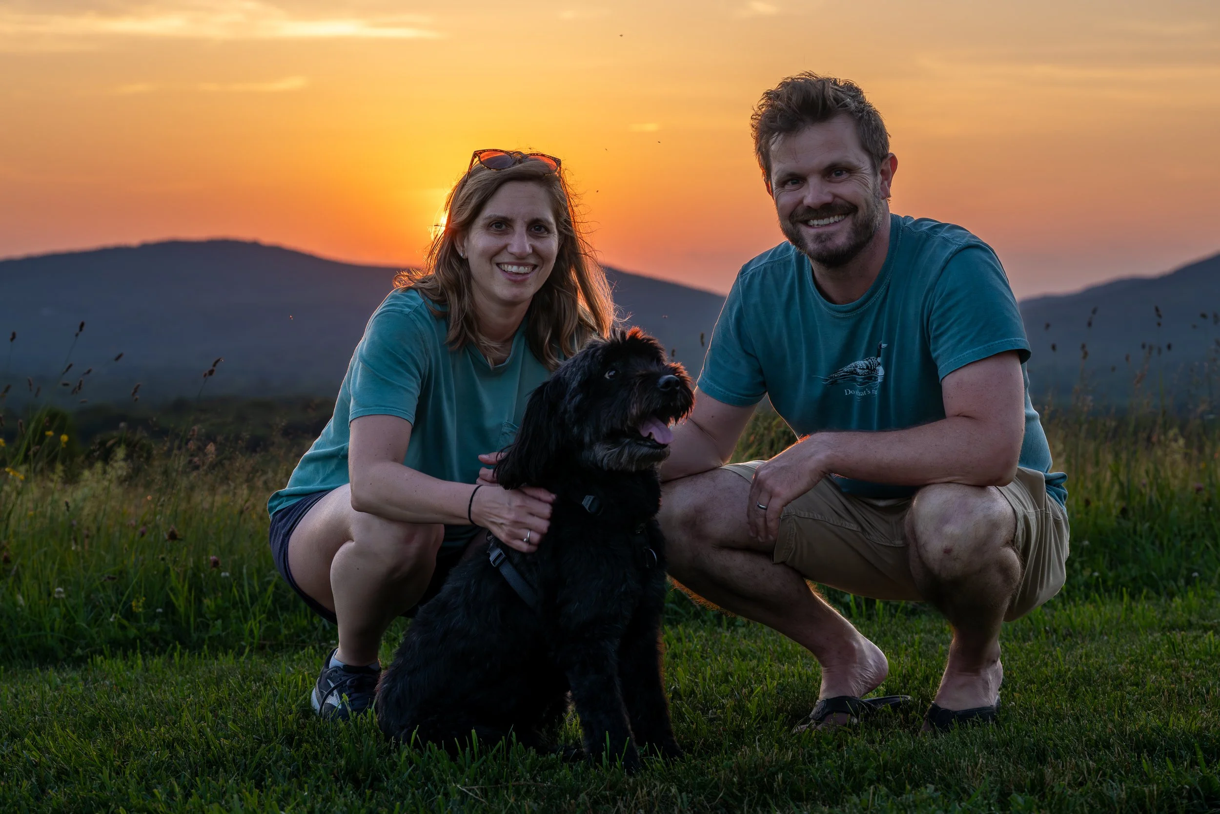 A smiling couple sitting on the grass with a black dog during sunset, mountains in the background, and orange sky.