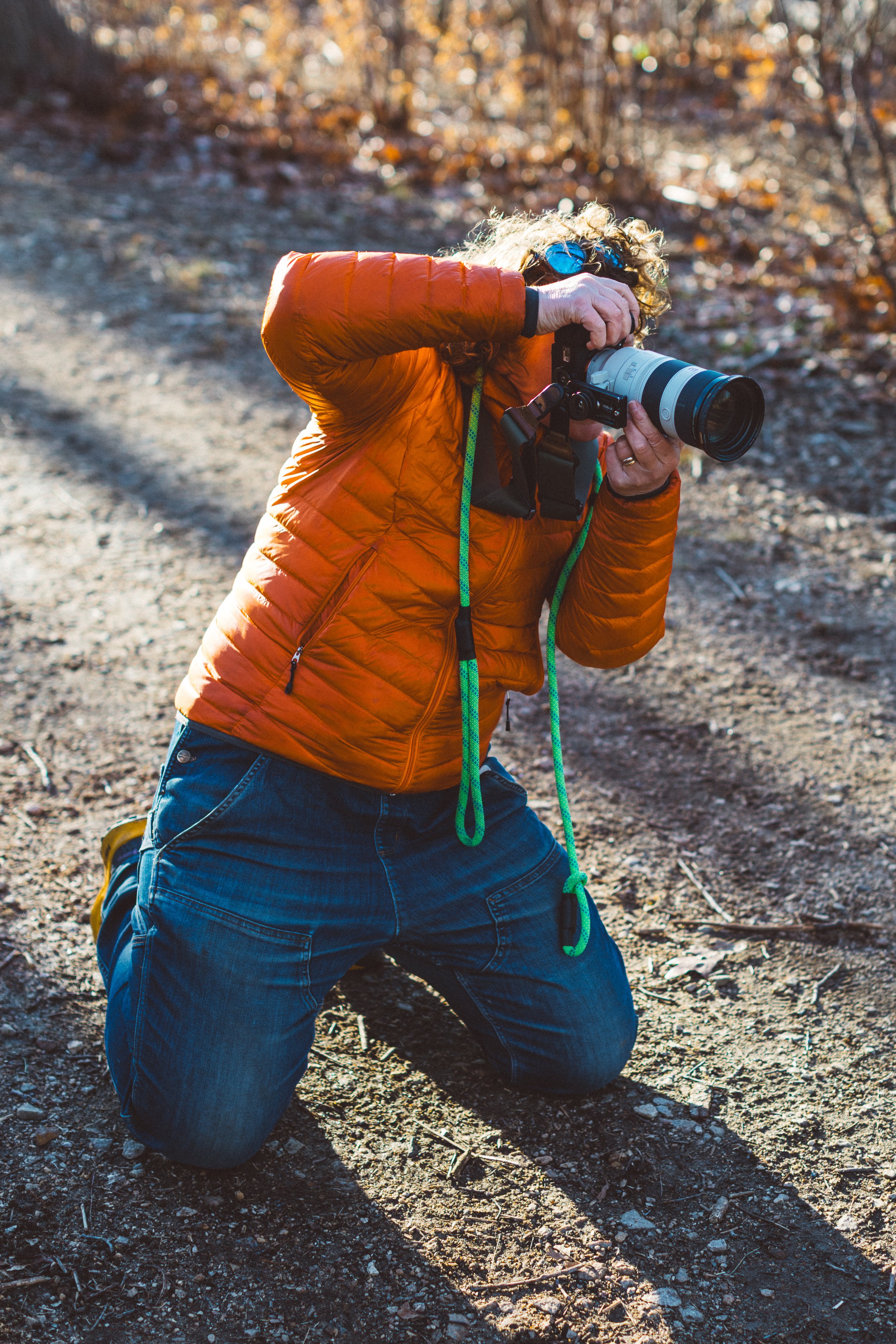 Jana in an orange jacket and jeans kneeling on dirt ground, taking a photograph with a large camera with a telephoto lens, outdoors in a wooded area with fall foliage. dog pet photographer