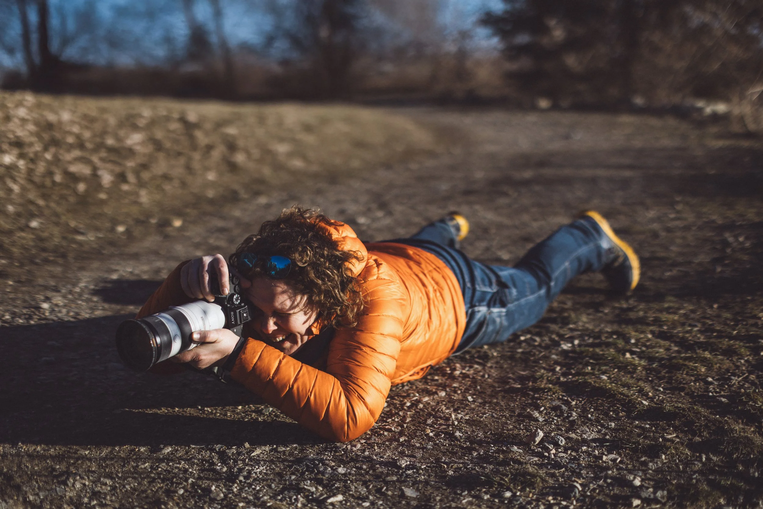Jana lying on the ground outdoors, taking a photograph with a camera, wearing an orange jacket, blue jeans, and yellow shoes. Dog Pet photographer New Hampshire new england