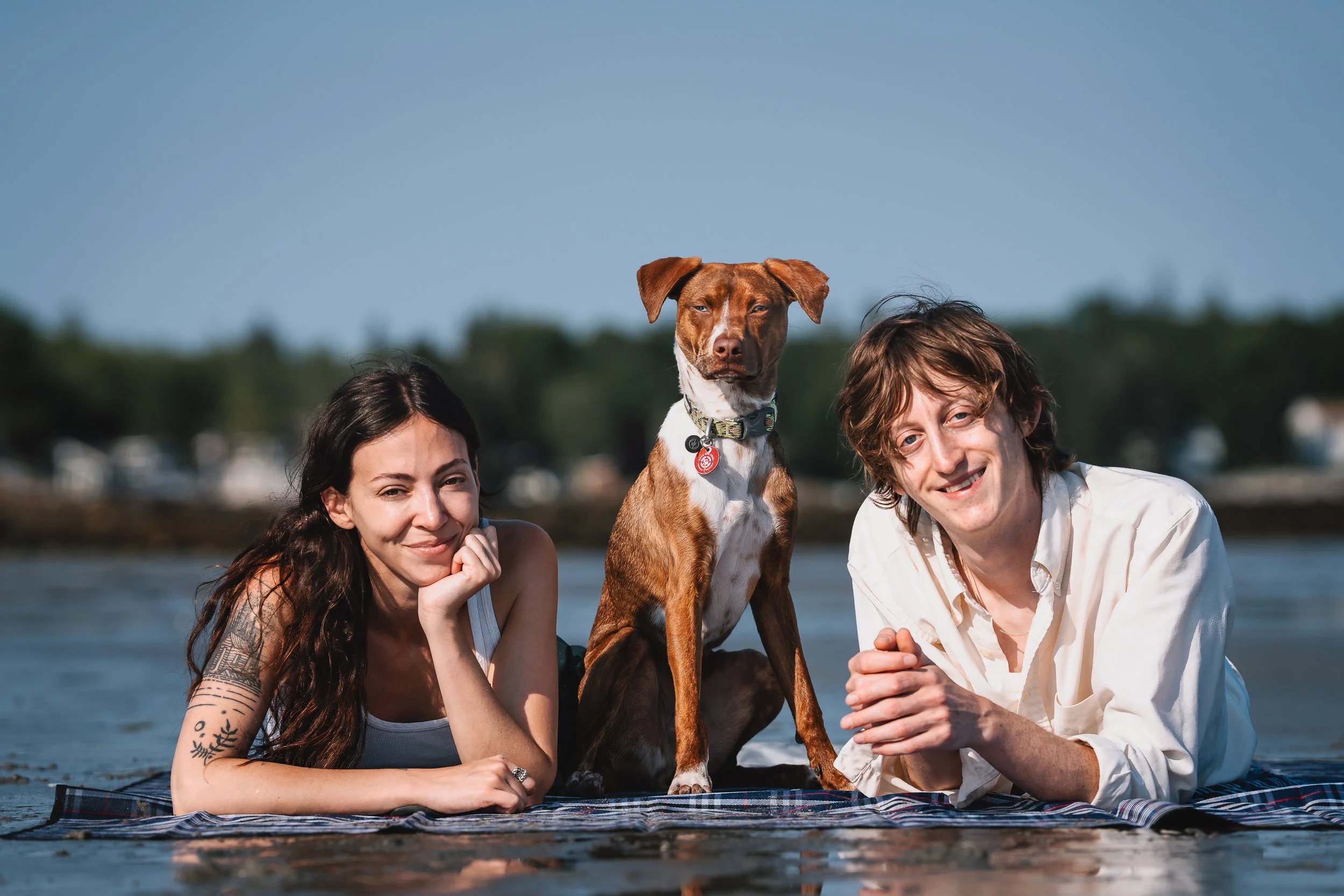 Two young adults, a woman with long dark hair and tattoos on her arm, and a man with curly brown hair, lie on a blanket at the edge of a body of water. Between them stands a brown and white dog with a red collar, looking directly at the camera. The b