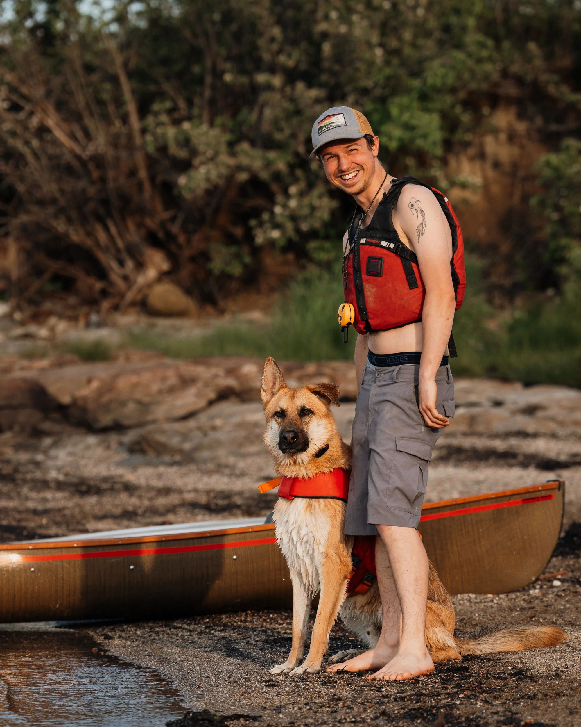 A smiling man in outdoor gear standing barefoot next to a dog on a beach with a canoe in the background. rockland Maine Midcoast maine Pet dog photography