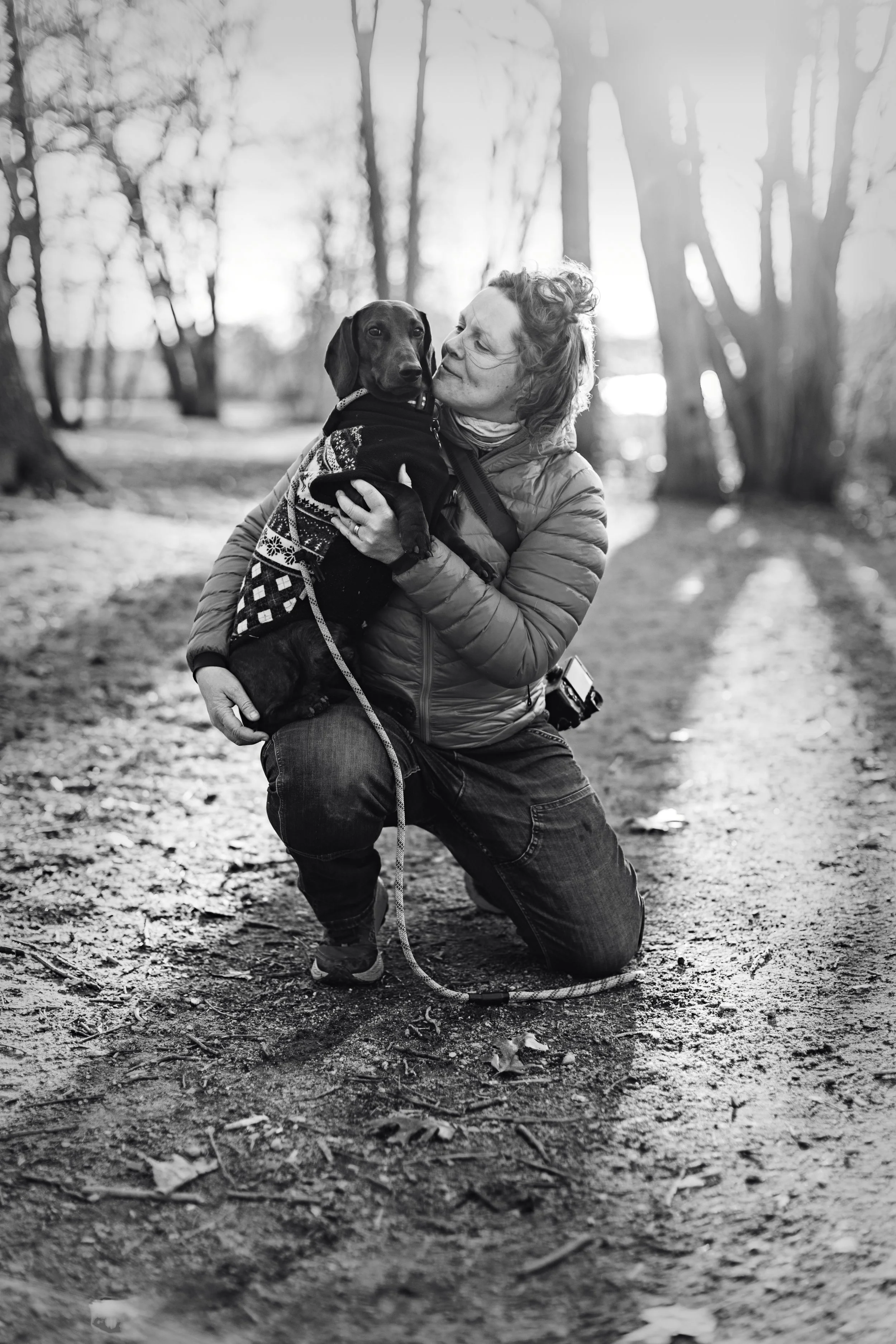 A woman kneeling on the ground outdoors, holding a dog dressed in a sweater, in a wooded area during the daytime.