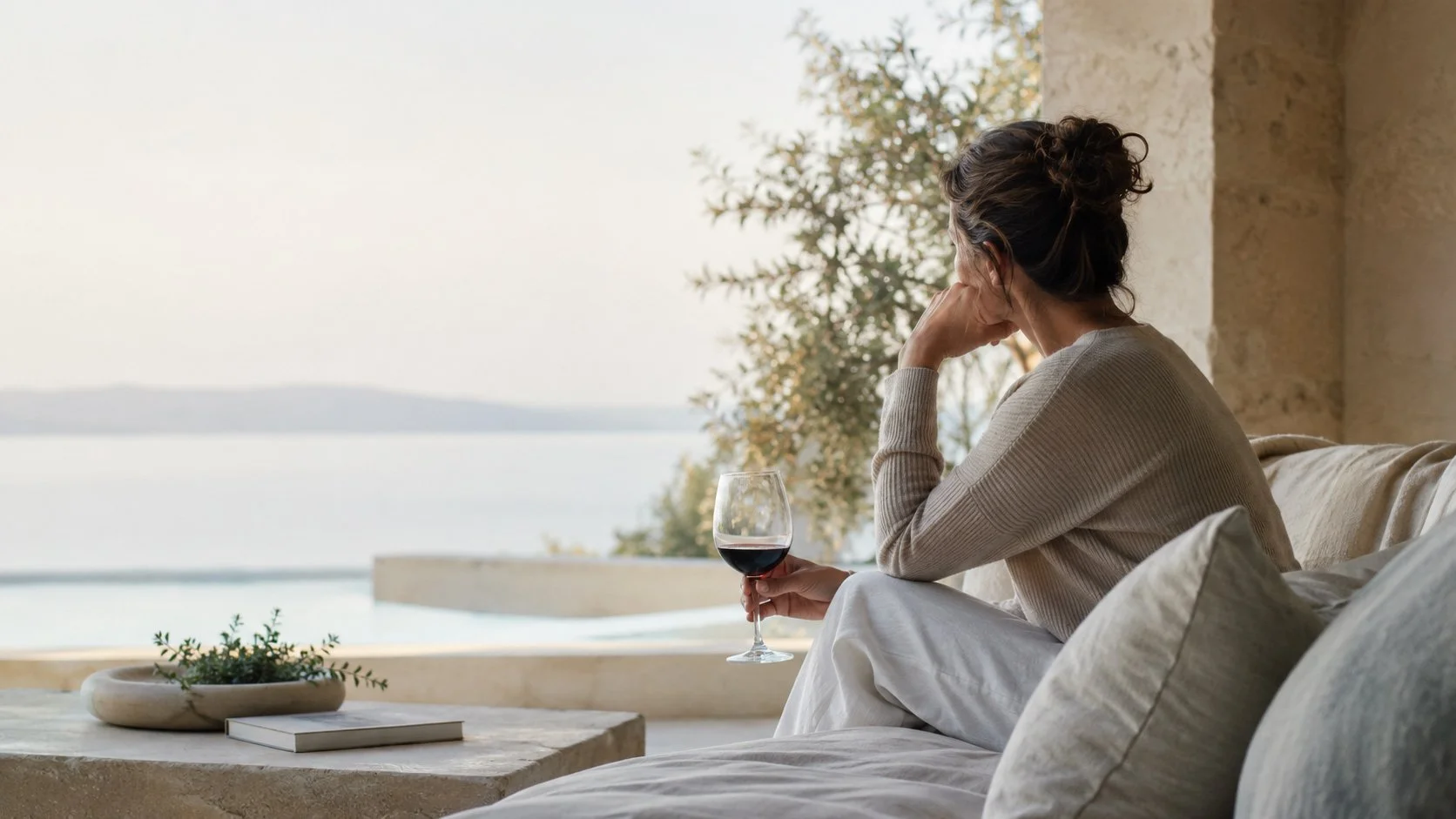 A woman with dark curly hair sitting on a sofa, holding a glass of red wine, looking out at a calm body of water with trees and land in the distance.