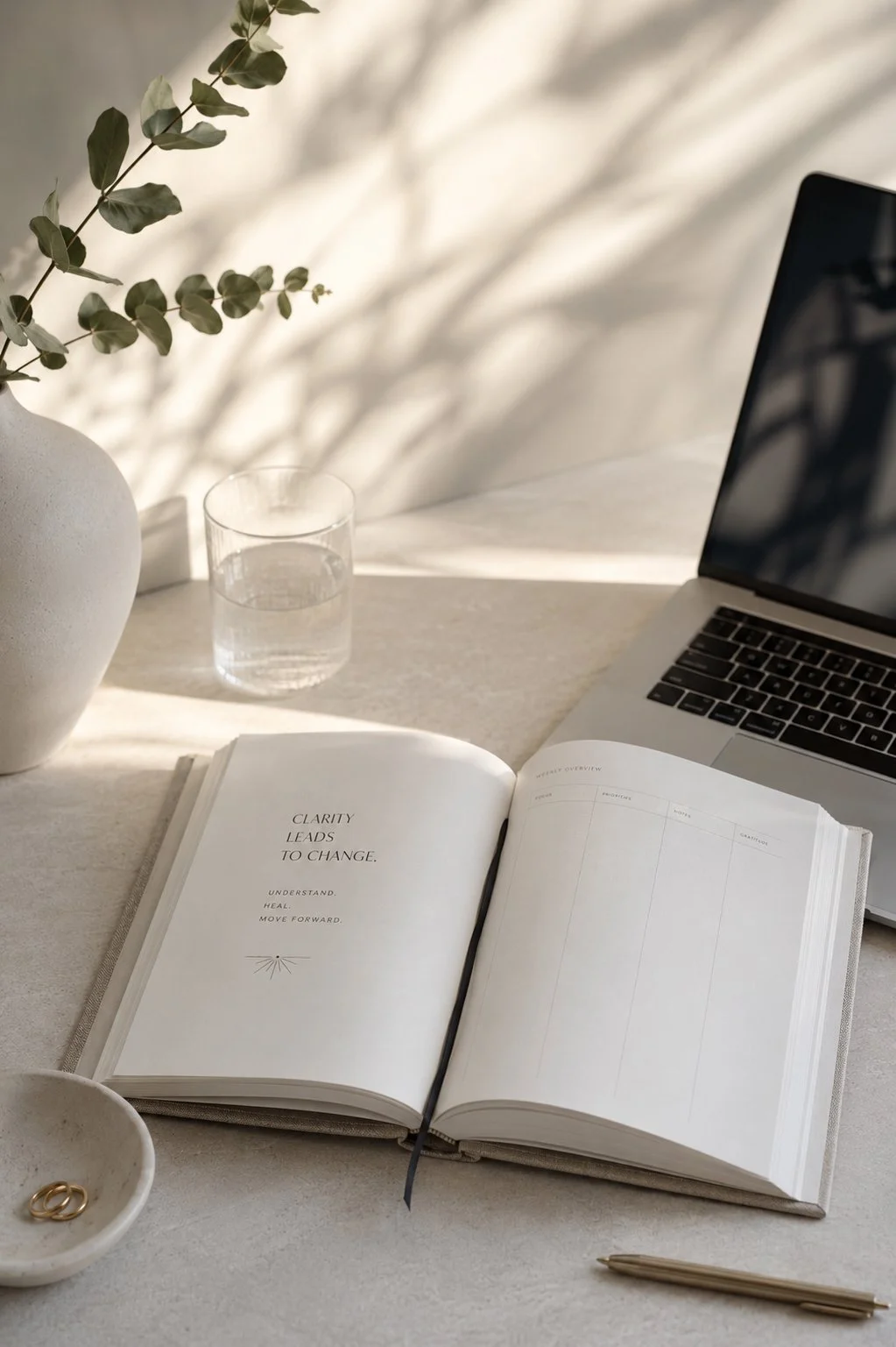Open journal with motivational quote, laptop, glass of water, potted plant, bowl with gold rings, and pen on a desk with sunlight and shadows.
