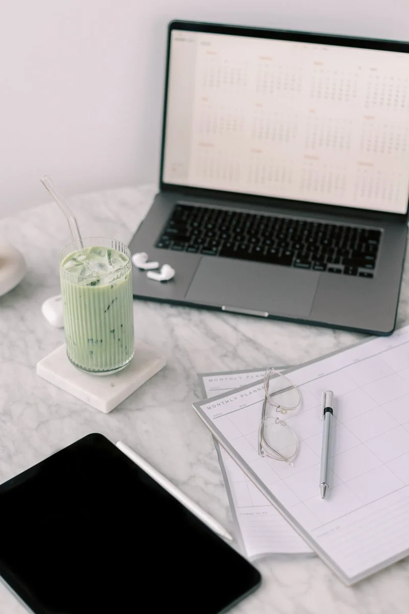 A work desk with a laptop displaying a calendar, a glass of green iced drink with a straw, a pair of glasses, a pen, a monthly planner, and a smartphone on a white marble surface.