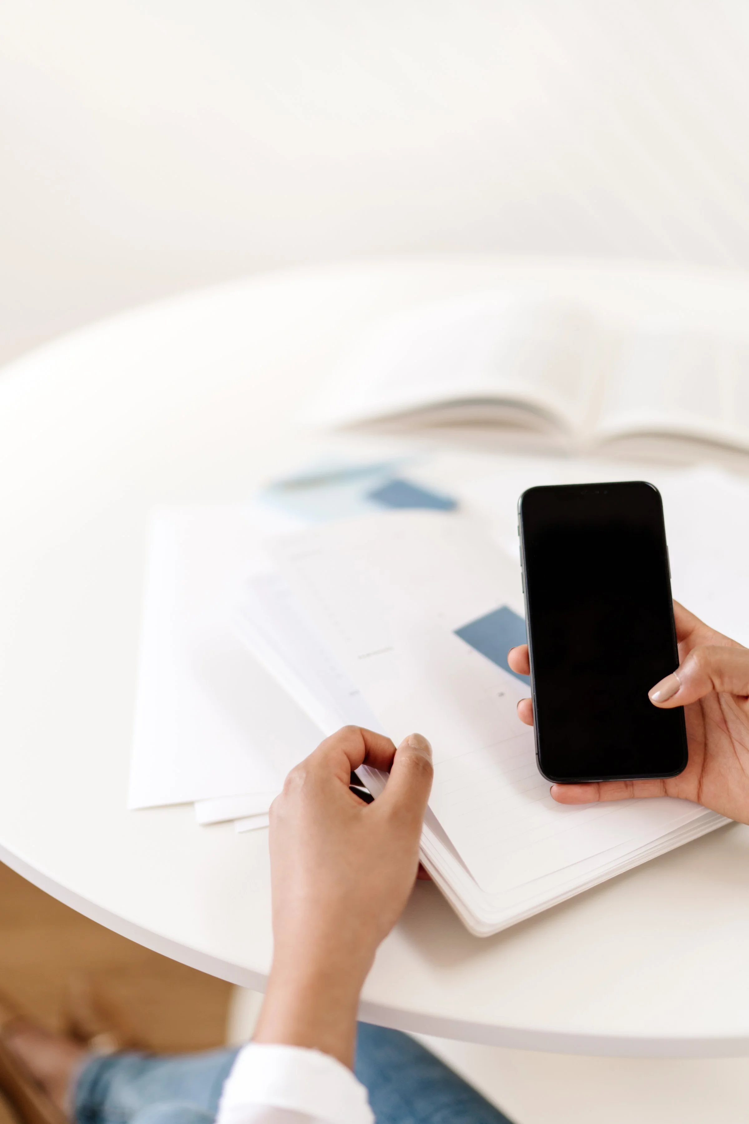 Person holding a smartphone in one hand and turning a page in a notebook with the other, on a white table with papers and an open book.