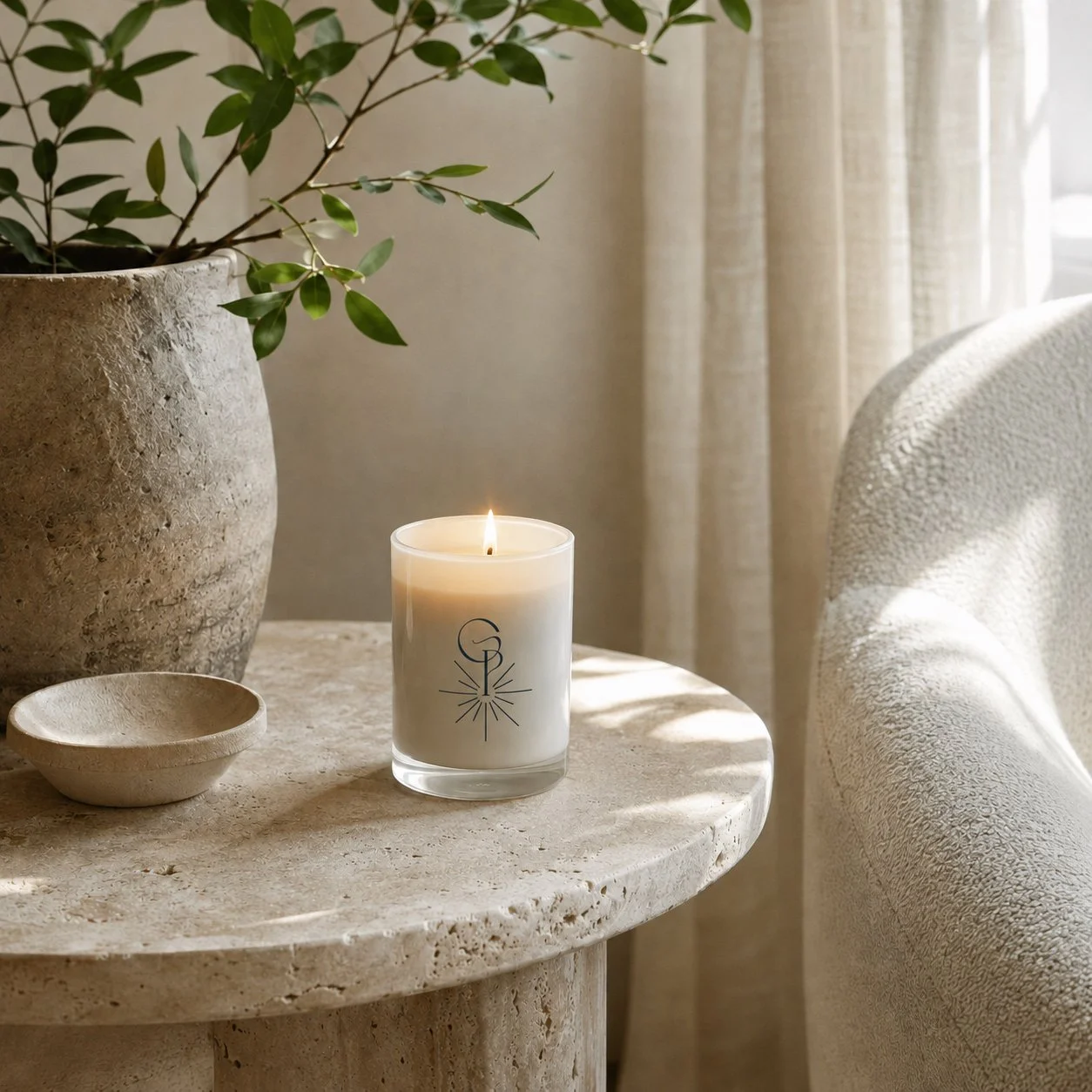 A lit white candle on a round stone table, with a potted plant and small dish nearby, in a cozy, well-lit room with beige curtains and a textured armchair.