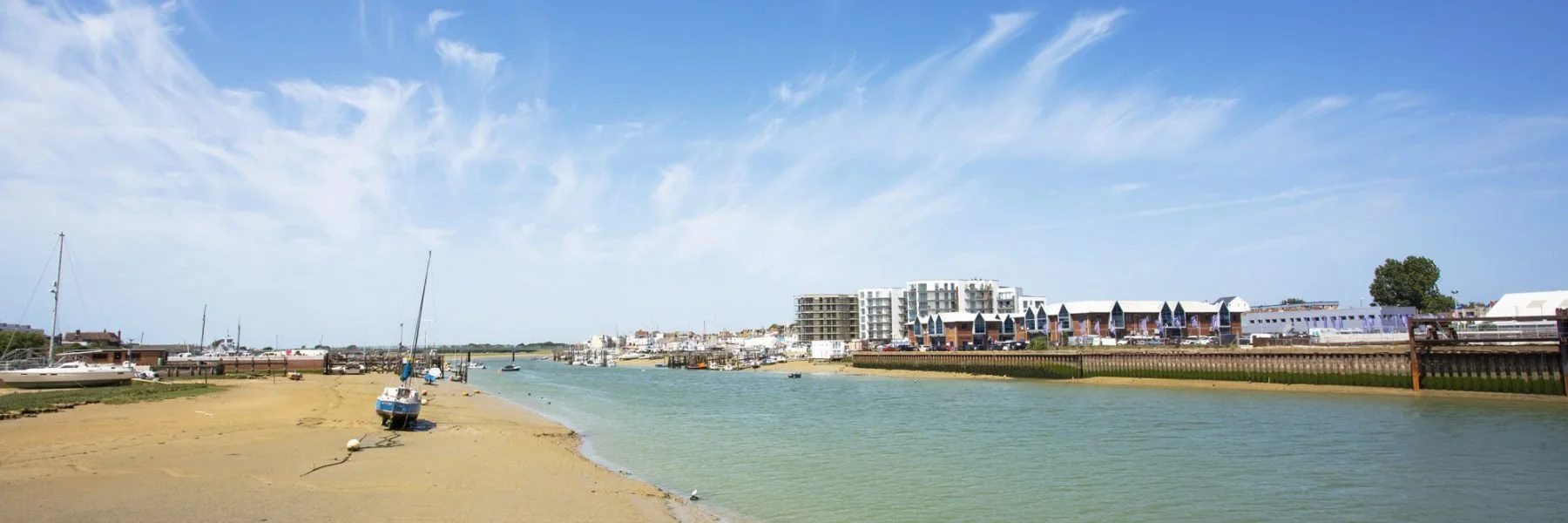 A coastal scene with boats anchored along a sandy shoreline, modern buildings in the background, and a partly cloudy sky.