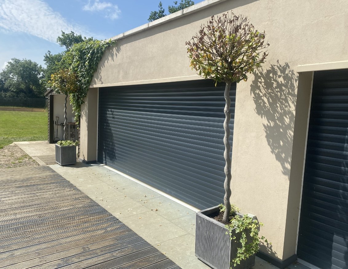 A gray building with a closed black metal garage door, two potted plants with small trees next to the door, and a grassy field in the background.