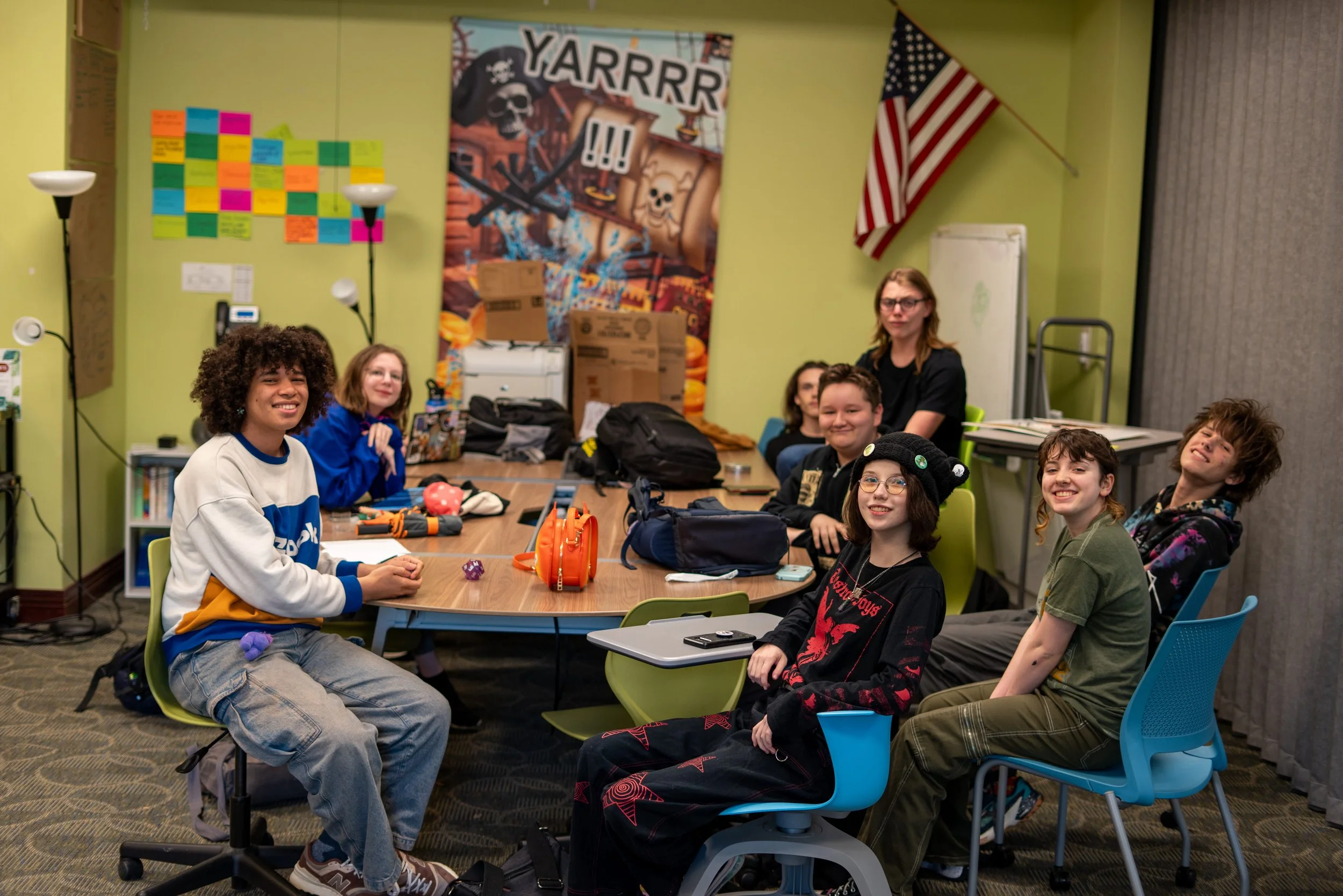 A group of young people and a woman sitting around a table in a colorful room with Green walls, a pirate-themed poster, and an American flag. They are smiling and appear happy.