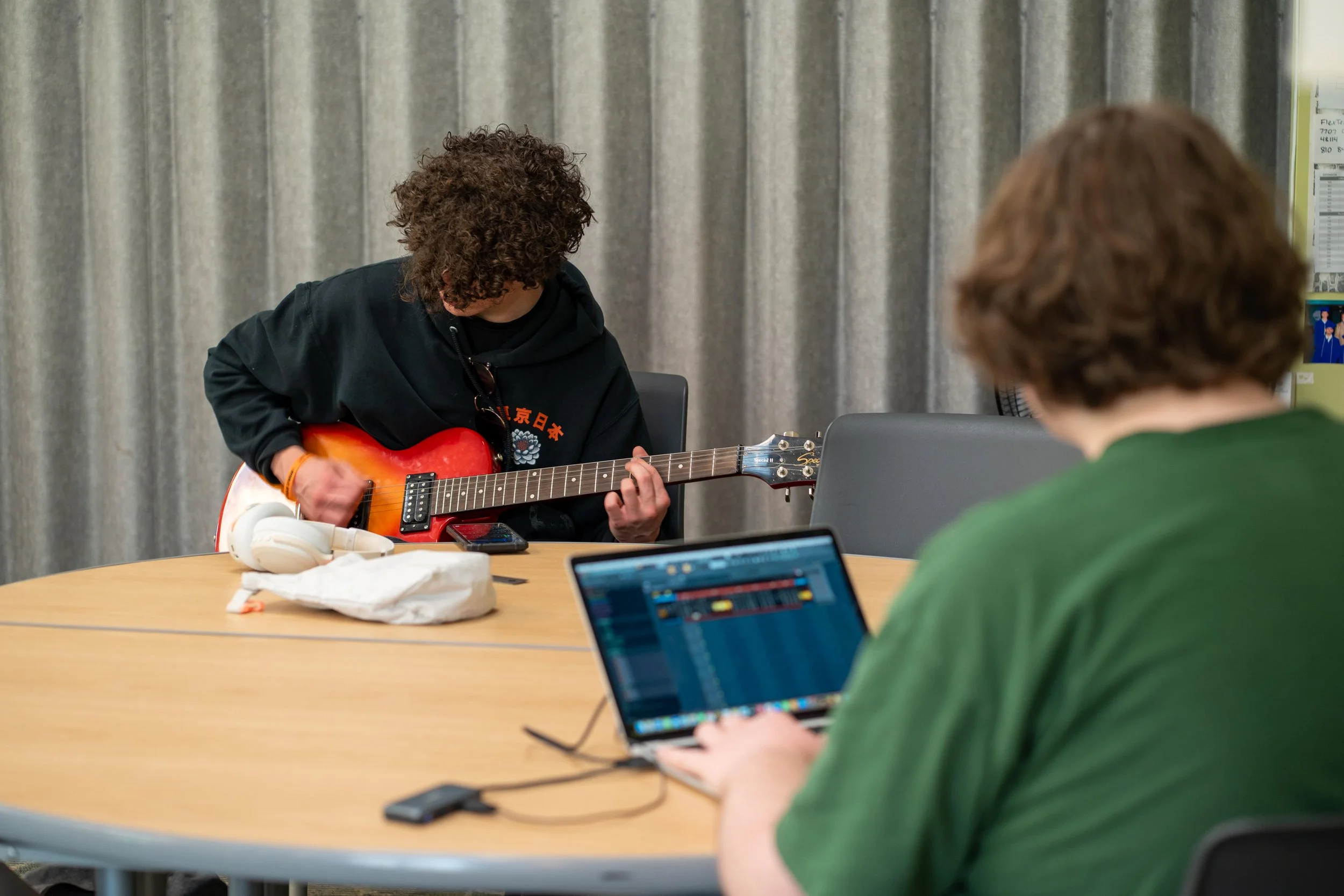 high school student playing an electric guitar while another person uses a laptop at a table at Brighton's alternative high school.