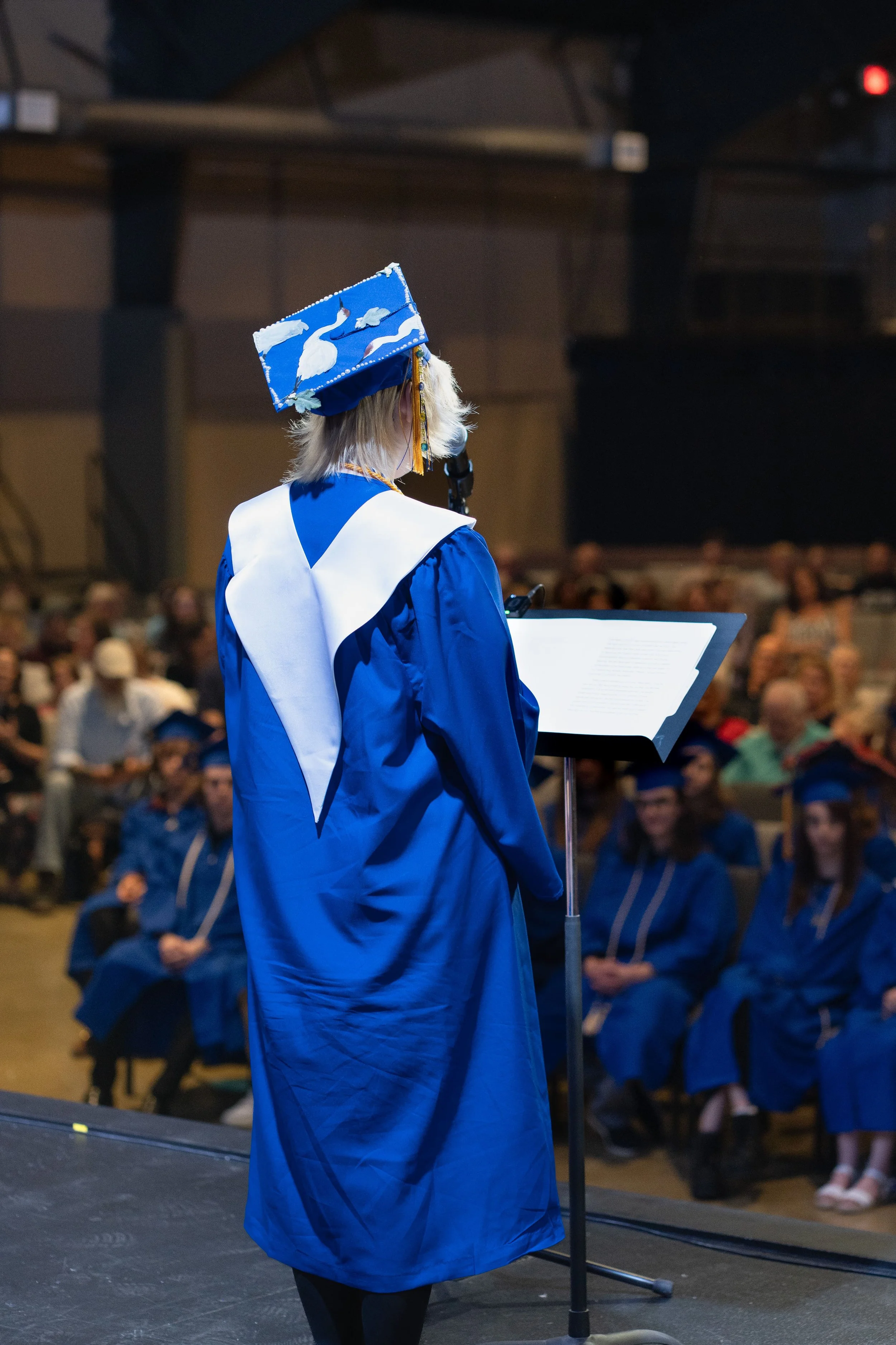 Graduation ceremony with a woman in blue robes and a mortarboard, reading from a podium, with an audience of graduates and spectators in the background.
