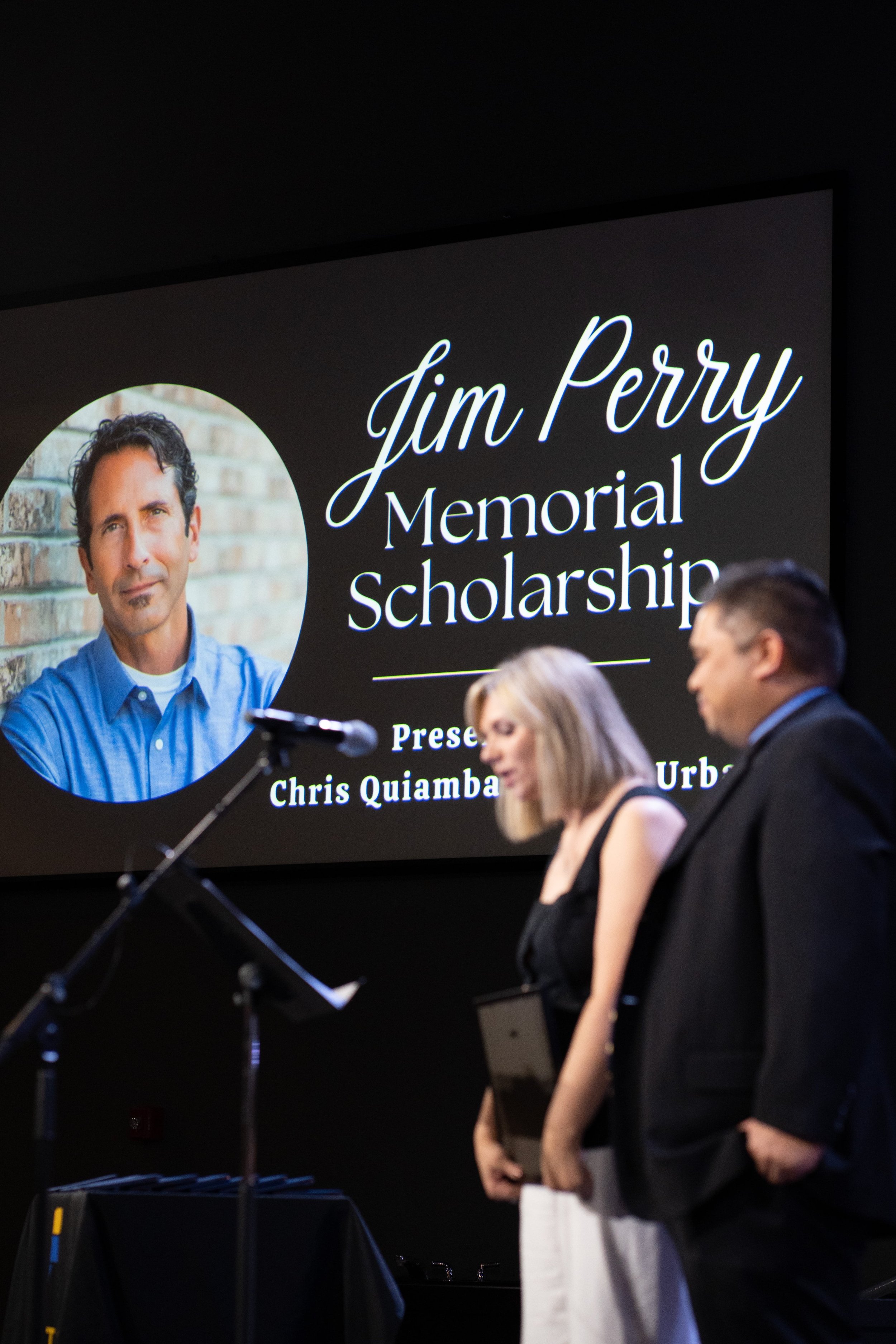 Presentation at the Jim Perry Memorial Scholarship event showing two people standing in front of a large screen with a photo of Jim Perry and event details.
