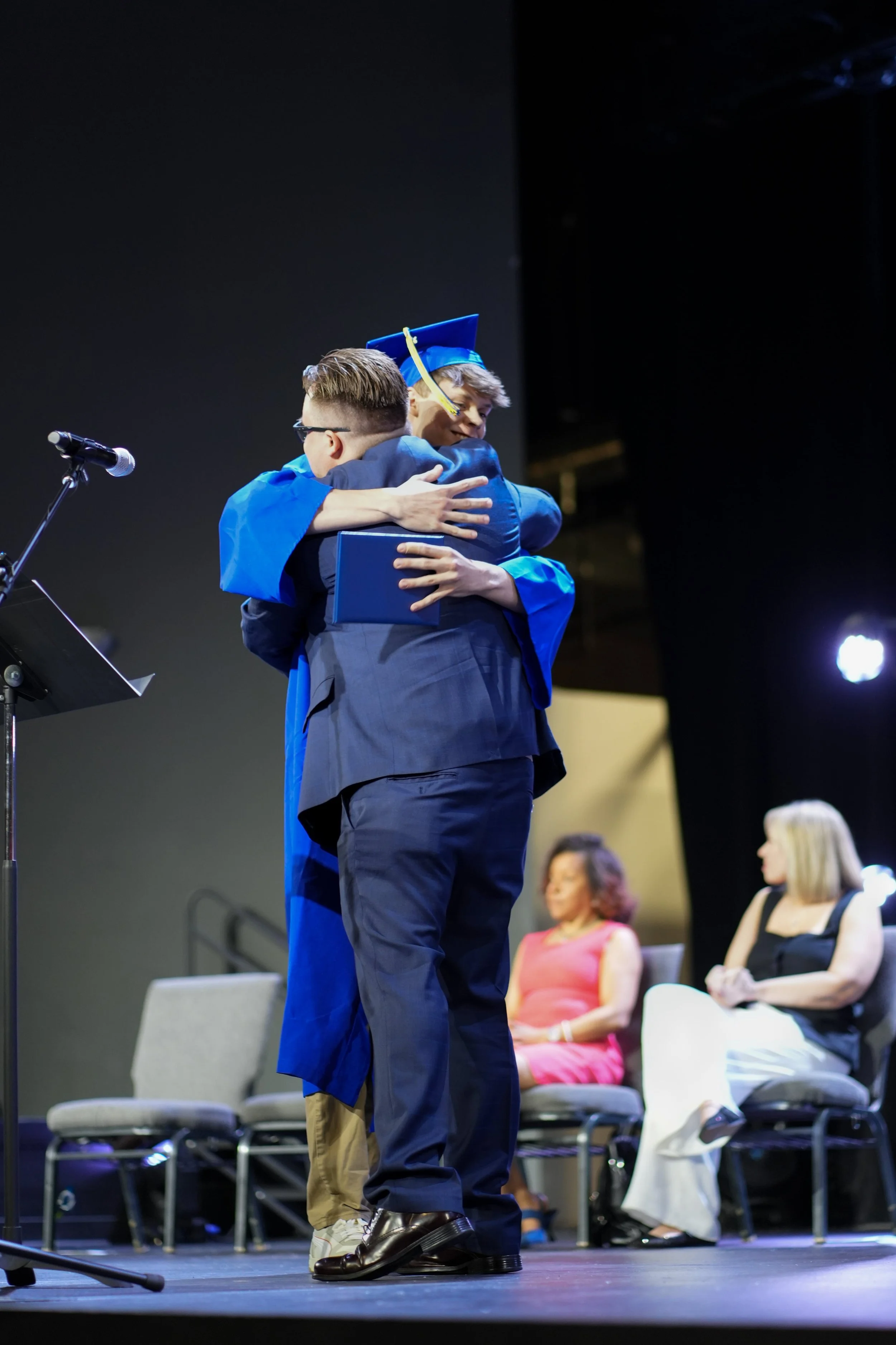Two people hugging on stage at FlexTech Brighton graduation ceremony, one in a blue cap and gown holding a diploma, with seated women in the background.