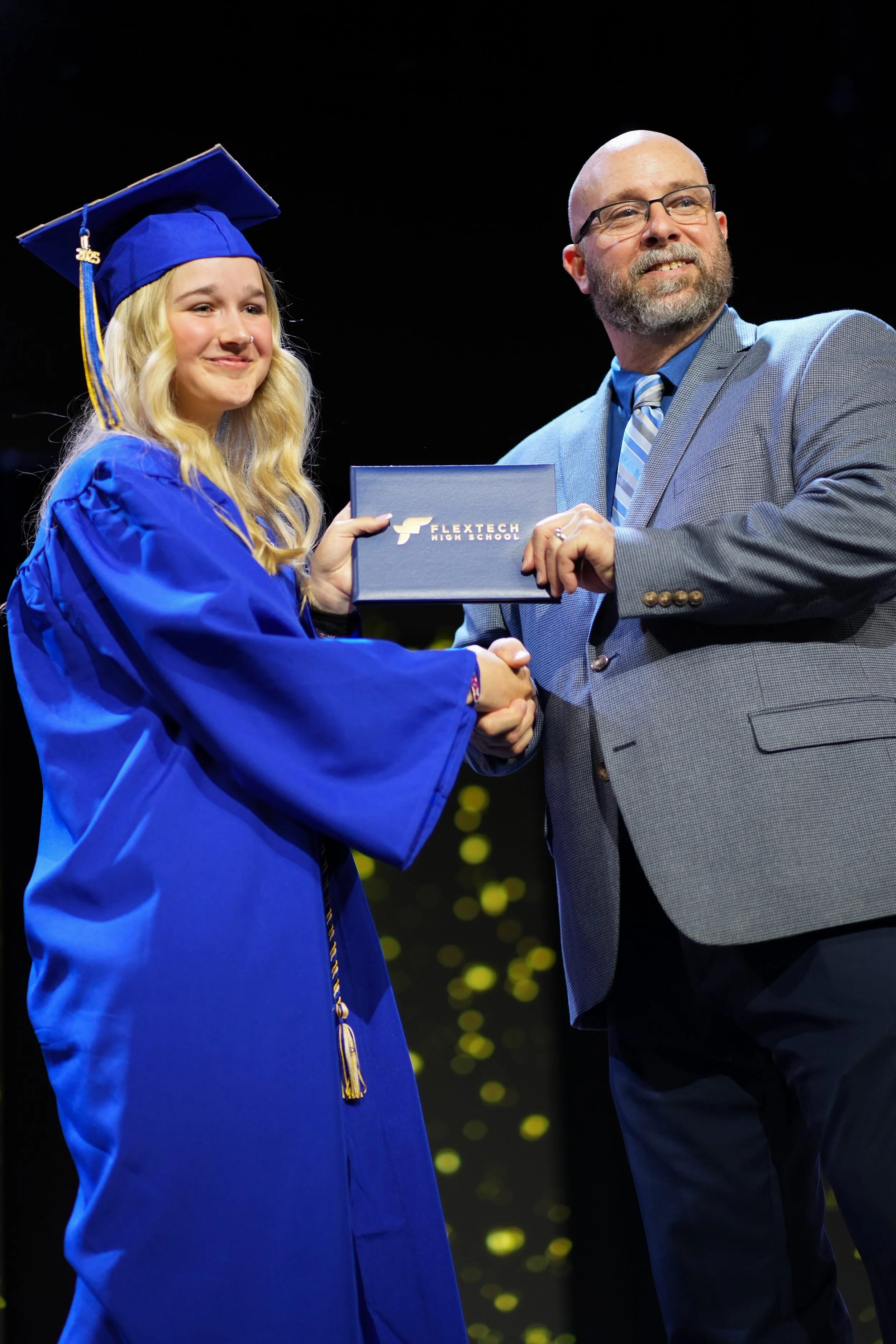 A young woman in a blue cap and gown shaking hands with a man in a gray suit during a graduation ceremony, holding a diploma from FlexTech High School.