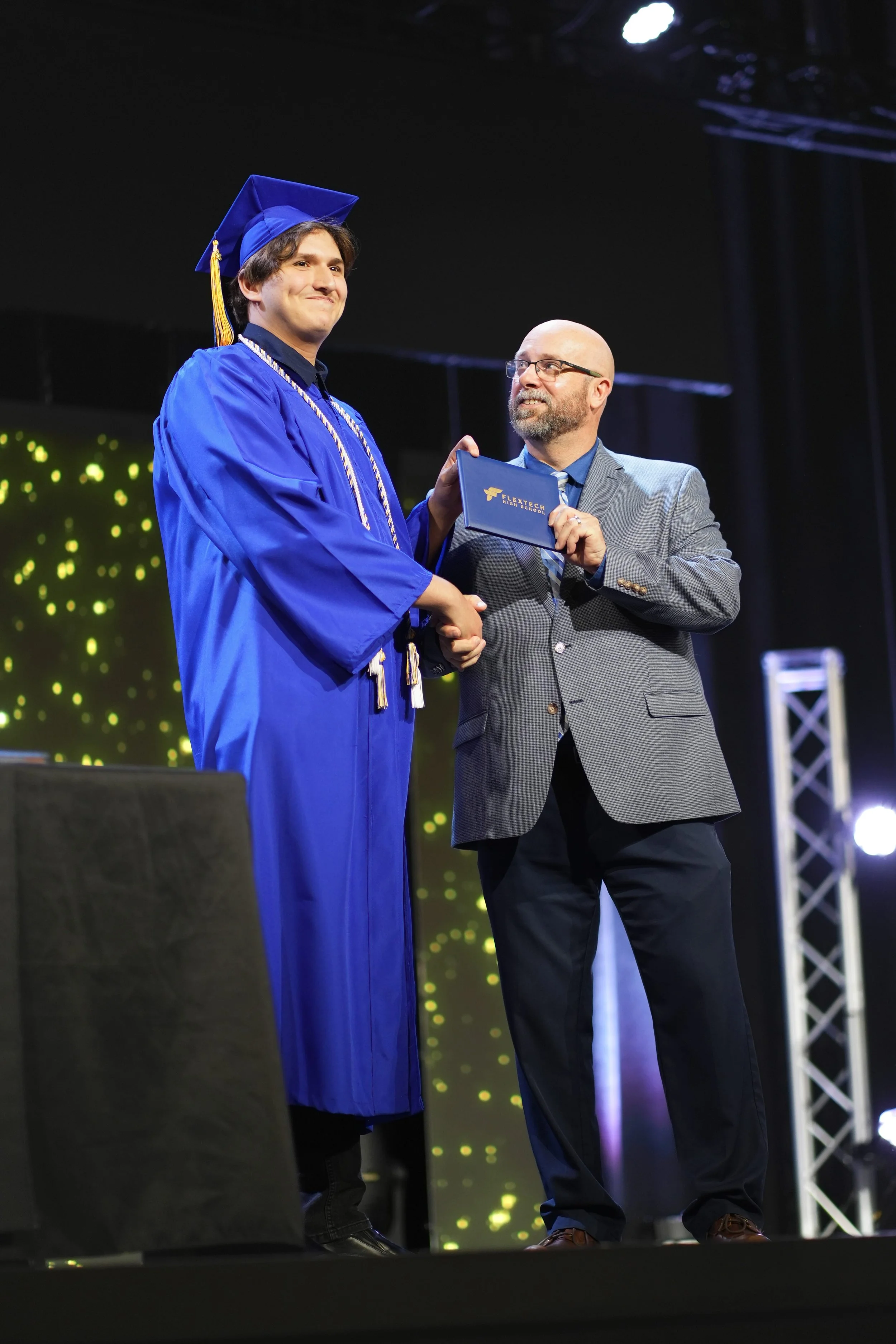 A young man in a blue graduation gown and cap shakes hands with Principal Jason, receiving a diploma or award.