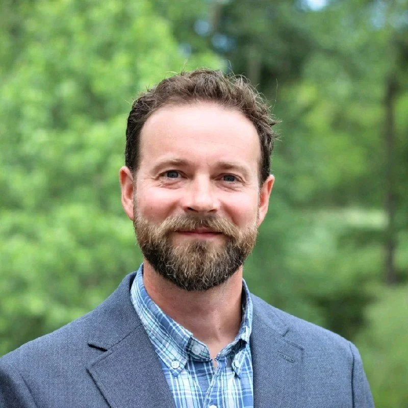 A high school teacher at FlexTech with glasses, long blonde hair, wearing a black blazer, standing with arms crossed against a gray background.